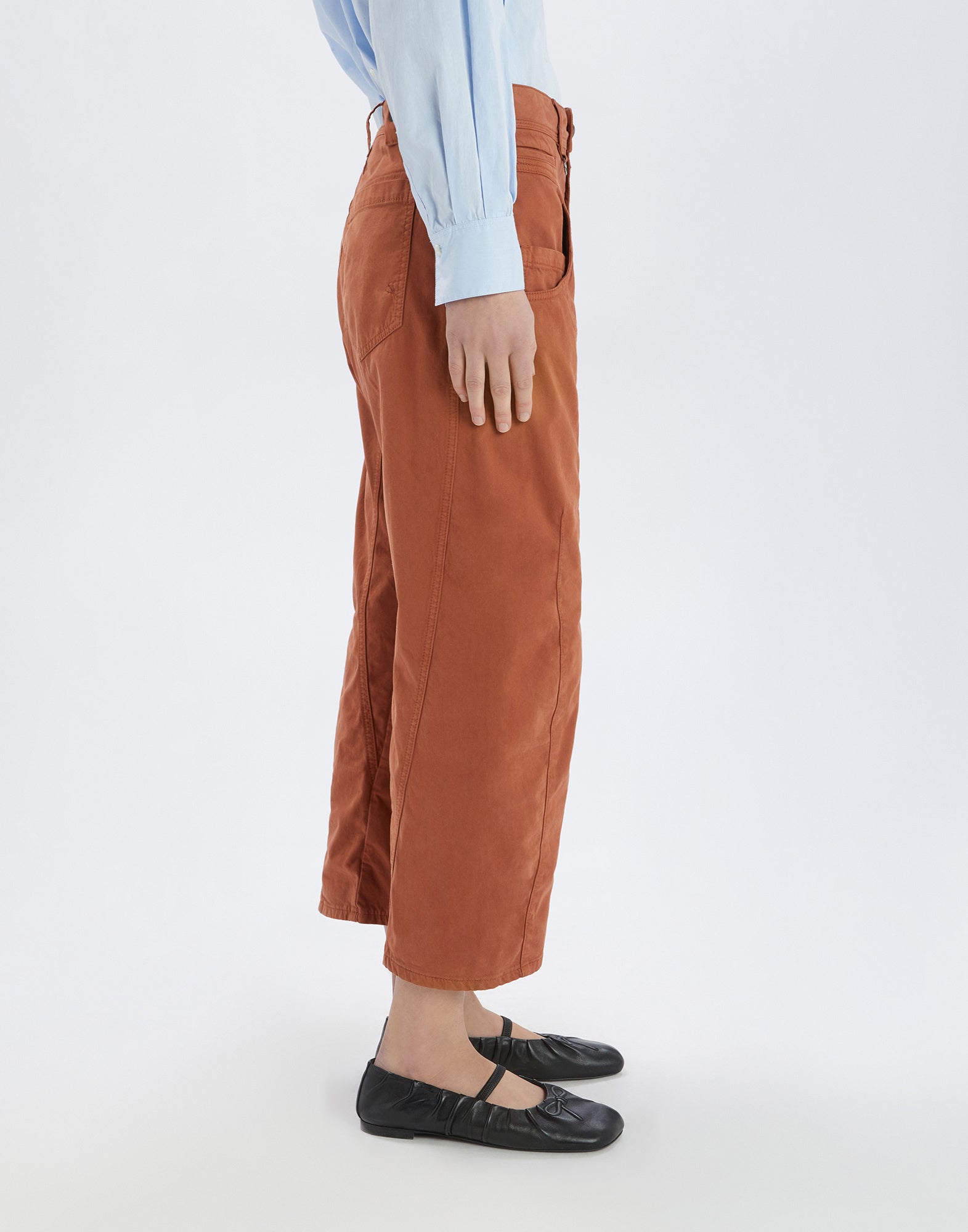 Seitenansicht einer Person, die die rostbraune Hose FURORE trägt, mit sichtbaren aufgesetzten Taschen und geschwungenen Nähten, kombiniert mit einem hellblauen Hemd.