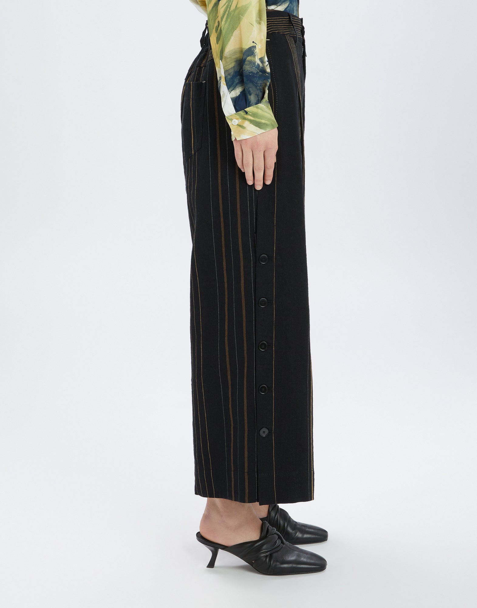 Seitenansicht der Hose SWALLOW mit vertikalen Streifen in verschiedenen Farben und Breiten auf schwarzem Grund, sichtbare seitliche Knopfleiste bis zum Knöchel.