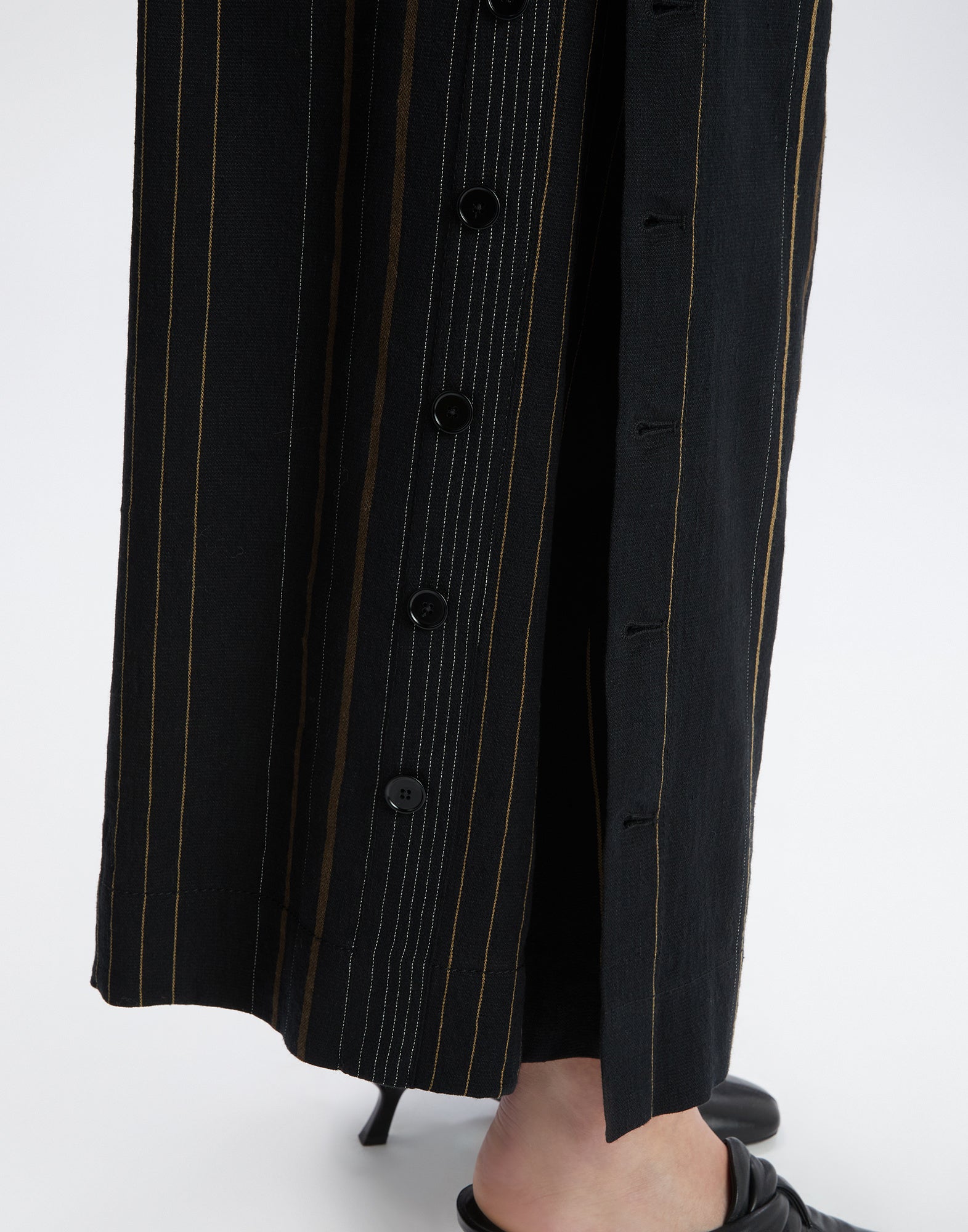 Detailansicht von SWALLOW Hose zeigt vertikale Streifen in verschiedenen Farben und Breiten auf schwarzem Stoff mit seitlicher Knopfleiste, getragen mit Absatzschuh.