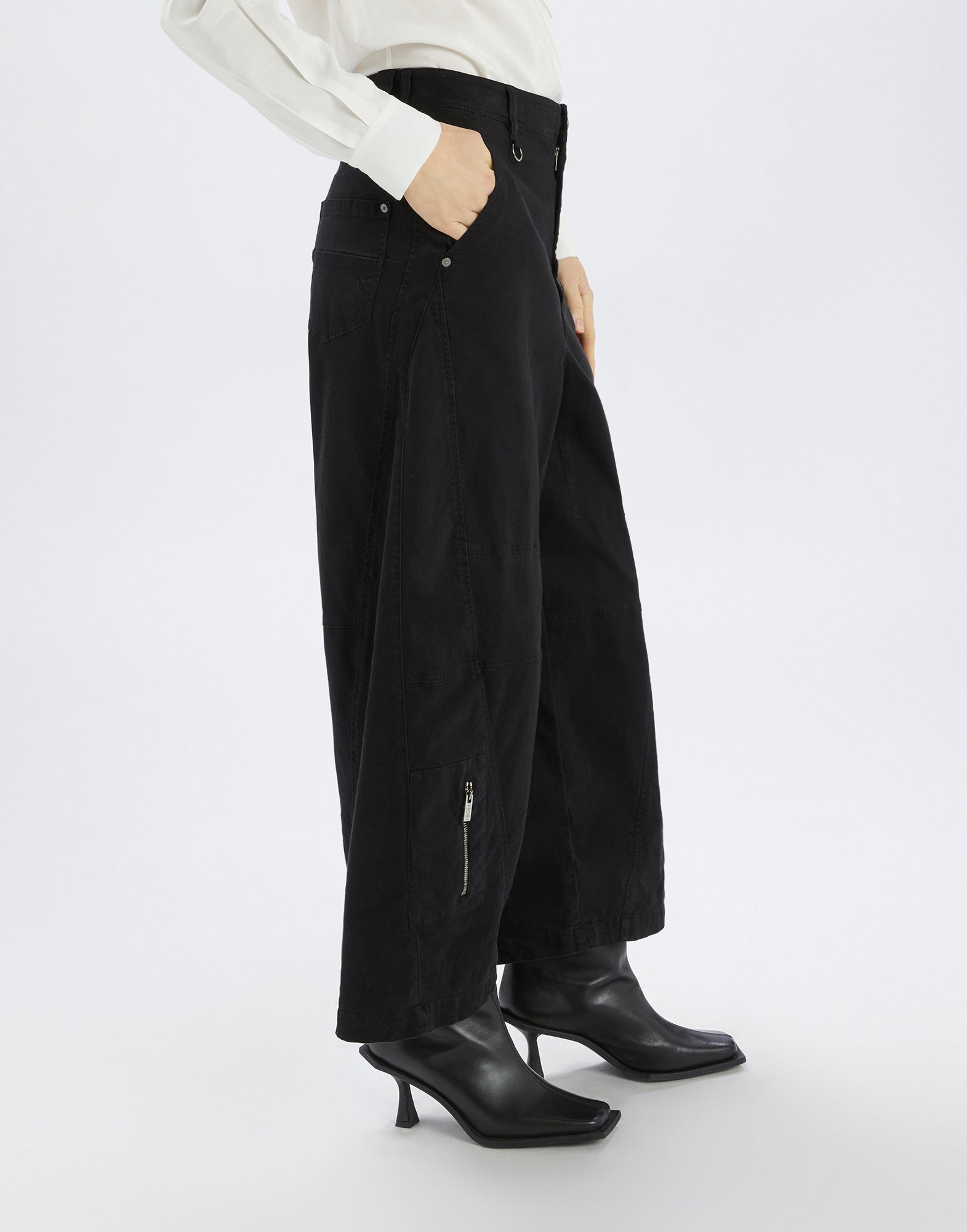 Seitenansicht einer Person, die die Hose OUT RUN trägt; schwarze, weit geschnittene Hose mit sichtbaren Steppnähten, vertikalen Reißverschlusstaschen und asymmetrischen Details.