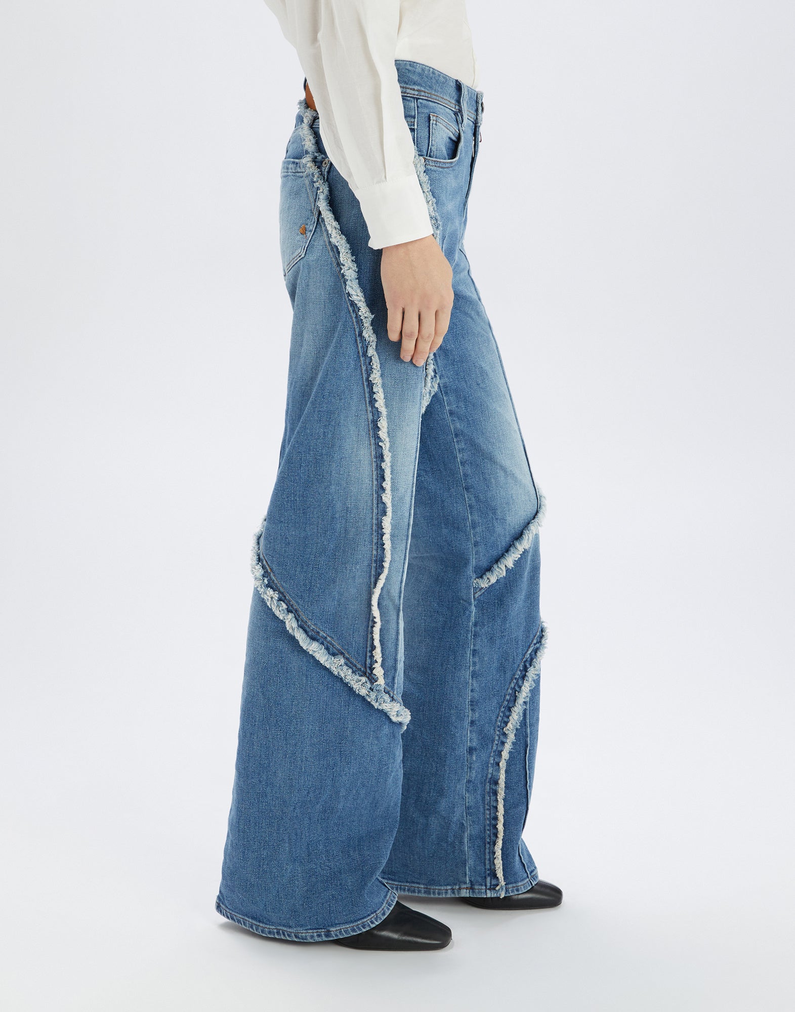 Seitenansicht der INDULGE Jeans mit weitem Bein, sichtbaren Außen- und Mittelnähten, offenen ausgefransten Kanten und hellblauer Waschung, getragen von einer Person.