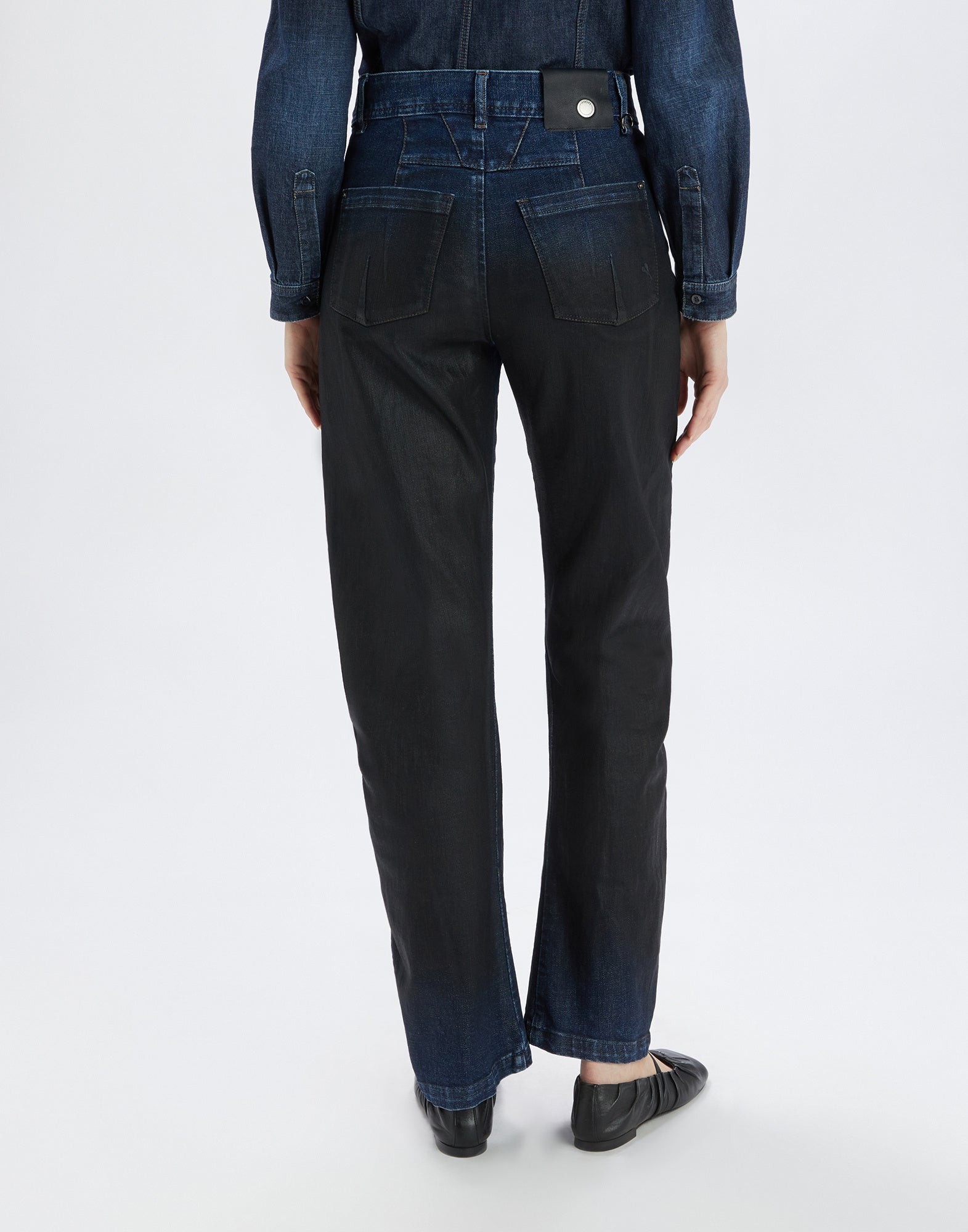 Rear view: Person trägt die TRICE Jeans, gerade geschnitten mit leicht gebogener Banana-Beinform, auffälligen Gesäßtaschen und sichtbaren Nähten im dunklen Denim-Look.