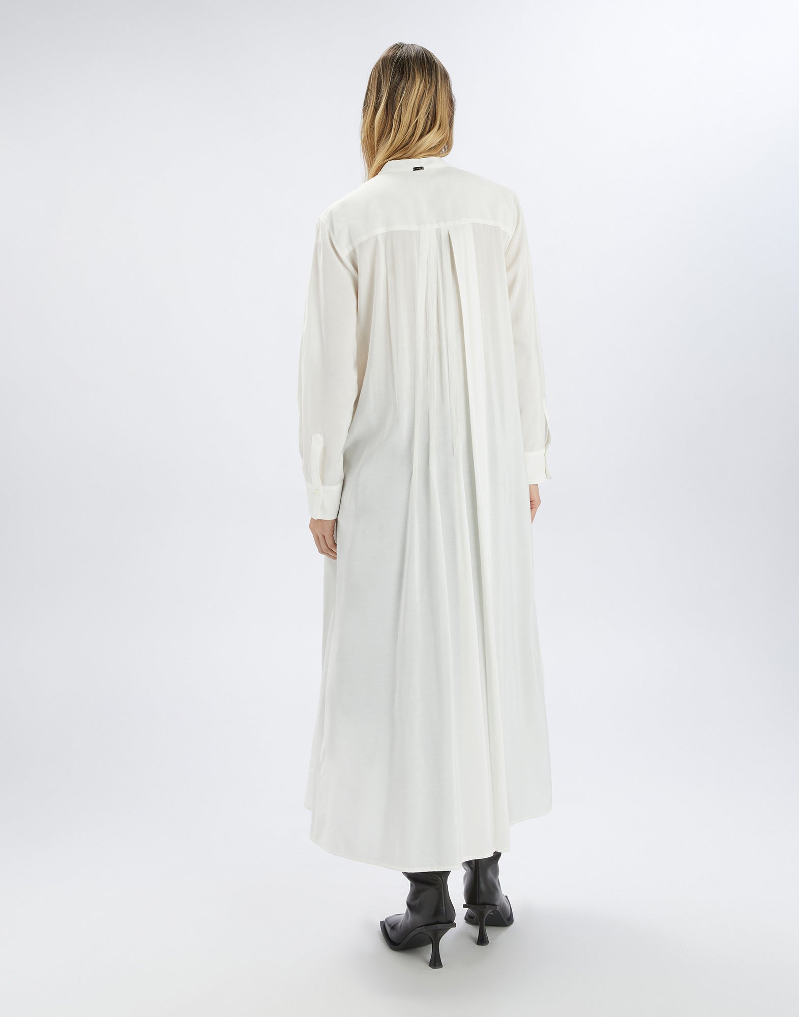Back view of the GALAXY shirt dress in white, featuring a long, loose fit, long sleeves, and irregular pleats, worn with black ankle boots.