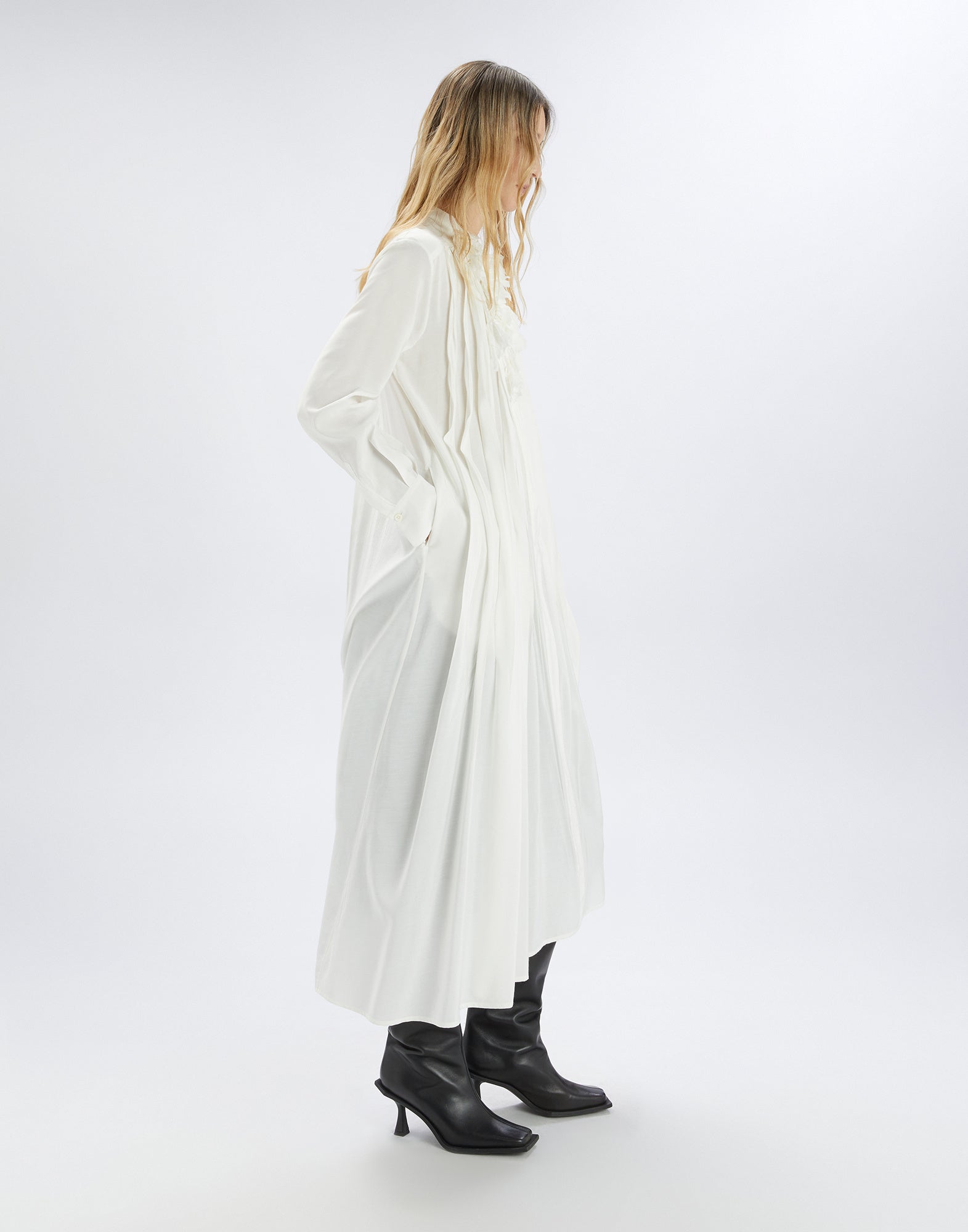 Side view of the GALAXY dress, white, with irregular pleats and floral mandarin collar detail, long sleeves and matching belt, styled with black boots.