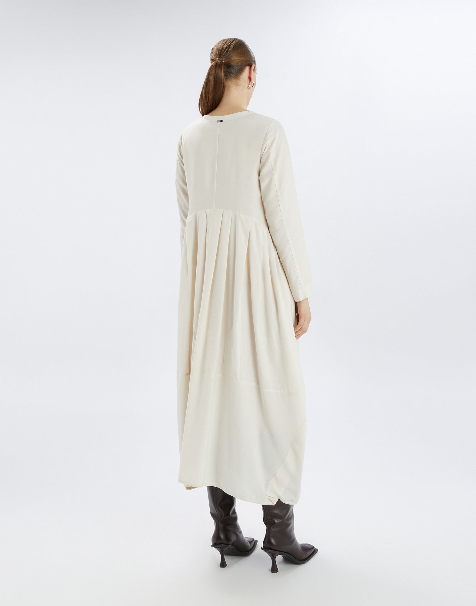 Back view of a person wearing the AWAKENING dress with long sleeves, rounded hem, and soft-flowing fabric, paired with dark boots.