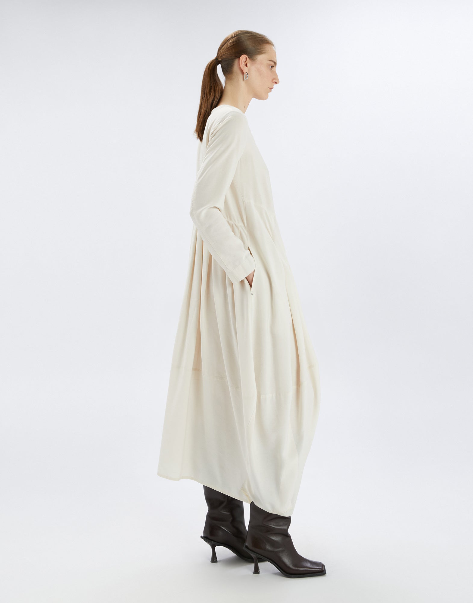 Side view of the AWAKENING dress, loose-fit with balloon skirt, long sleeves, and pockets, worn with dark boots against a light background.
