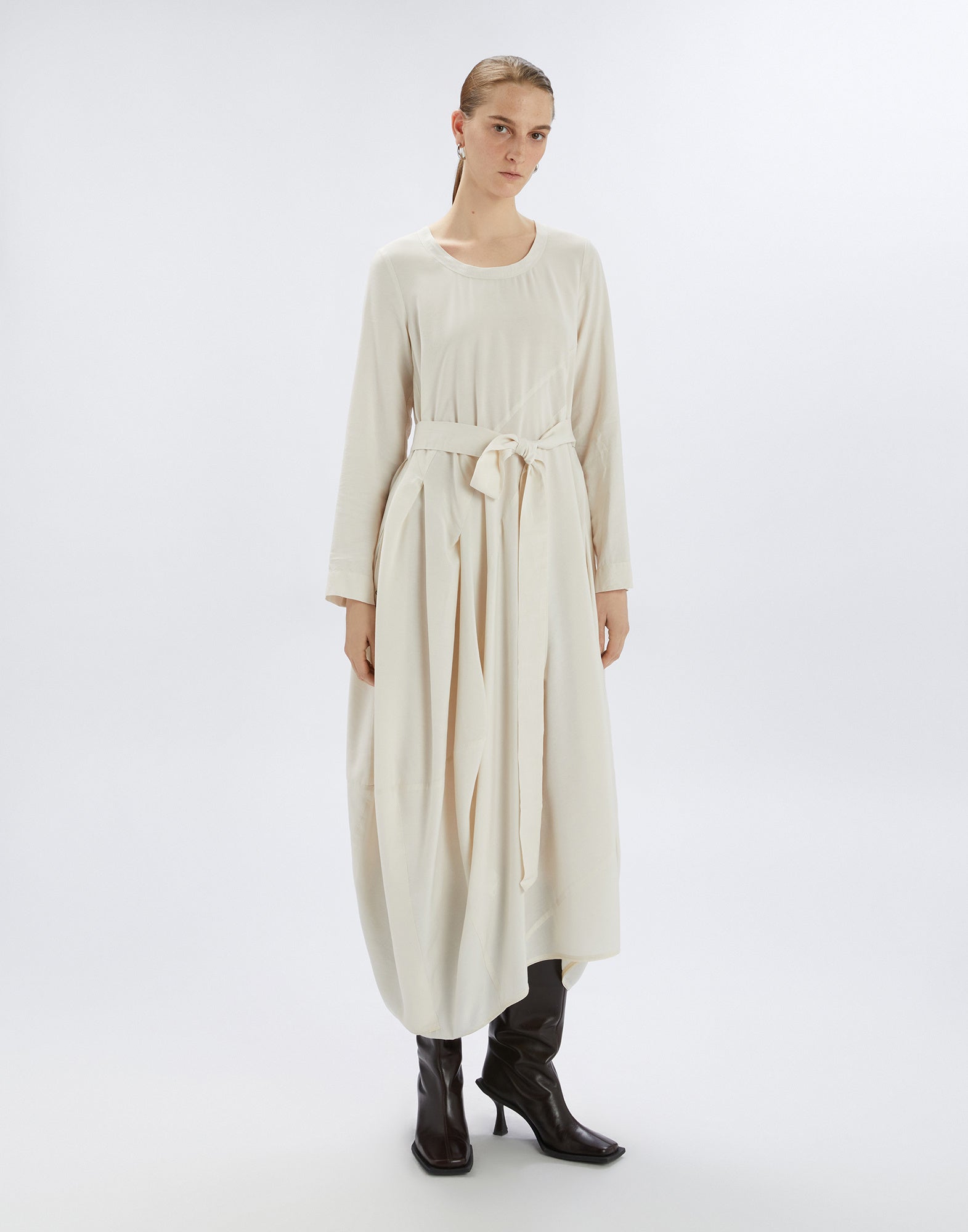 Front view of the AWAKENING dress, shown on a standing woman. The dress features long sleeves, round neckline, balloon skirt, waist belt, and asymmetric hem.