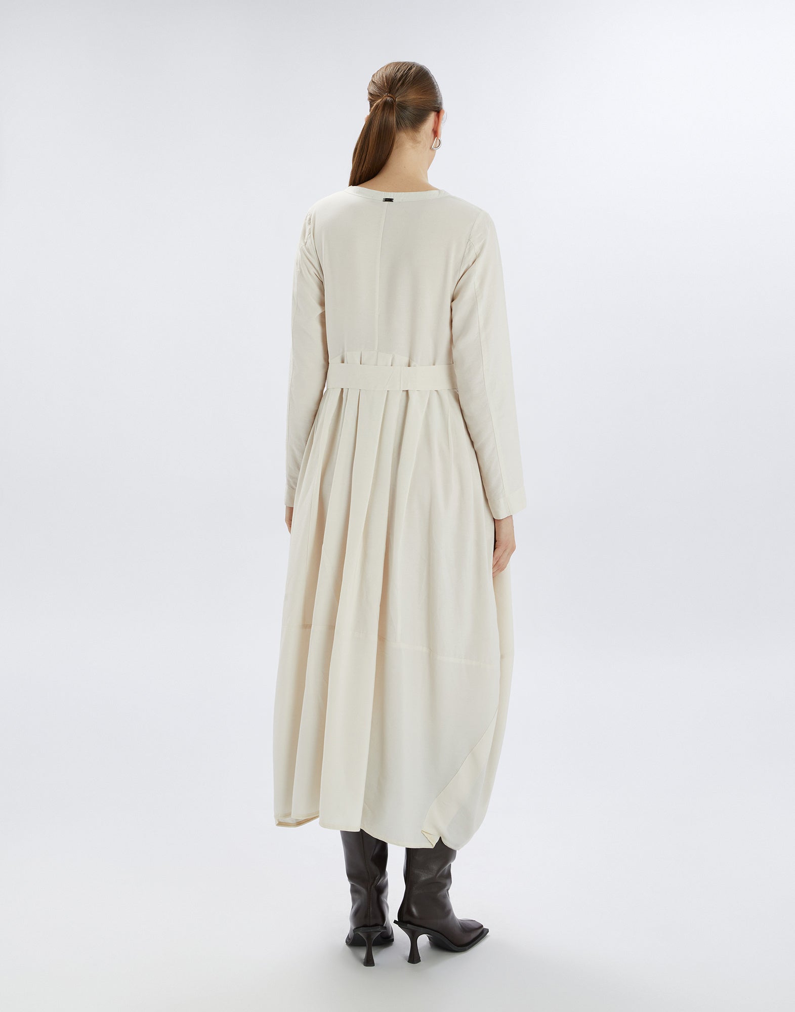 Back view of a woman wearing the AWAKENING dress; light-colored, long dress with balloon skirt, long sleeves, and fabric belt, paired with dark boots.