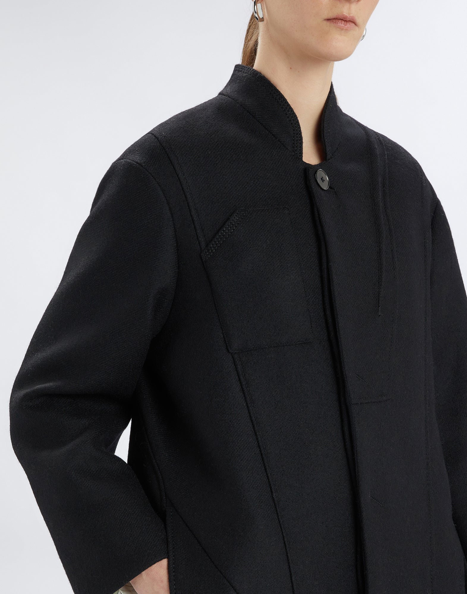 Detailansicht von vorne: Der schwarze PANTO Mantel mit Stehkragen, sichtbarem Knopfverschluss und seitlicher Nahttasche wird im Oversize-Stil präsentiert.