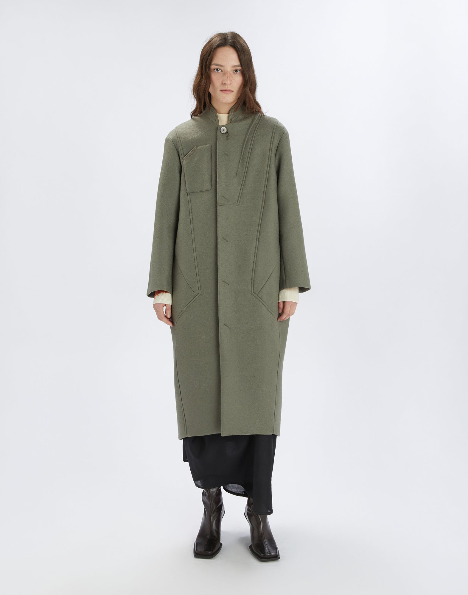 Front view: Woman wears the PANTO coat, ankle-length, oversized with stand collar, visible button, and chest pocket in olive green, against a neutral background.