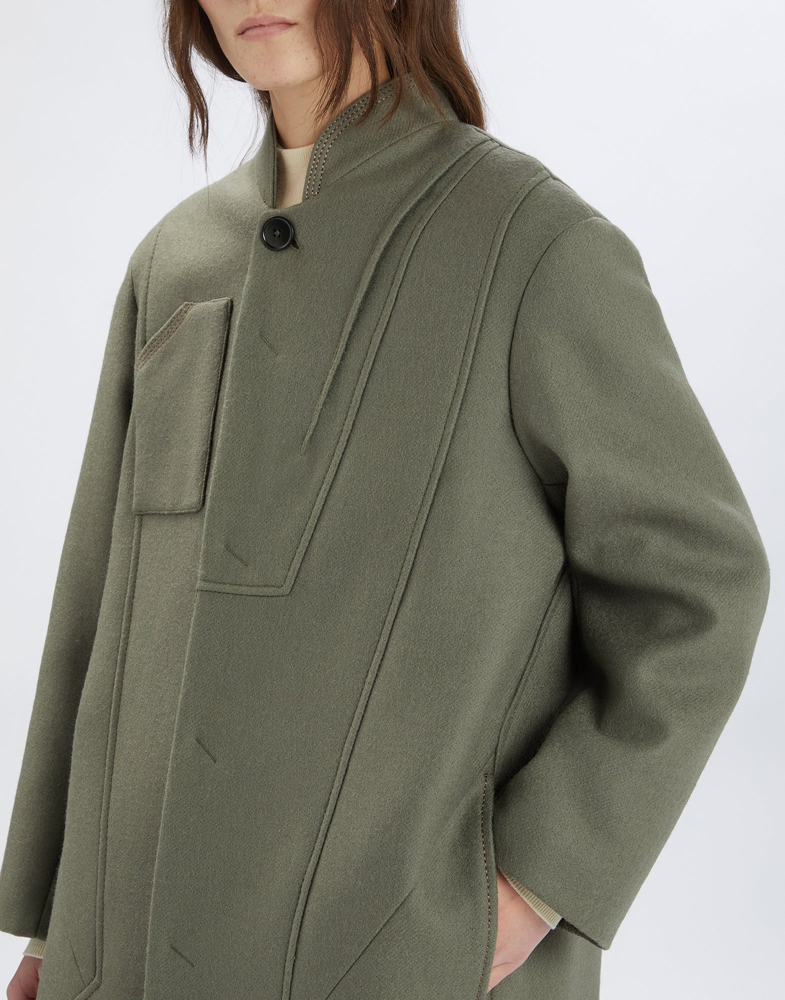 Detail view of the PANTO coat in black wool with oversized fit, stand collar, striking seam and visible button, worn by a person with hand in pocket.