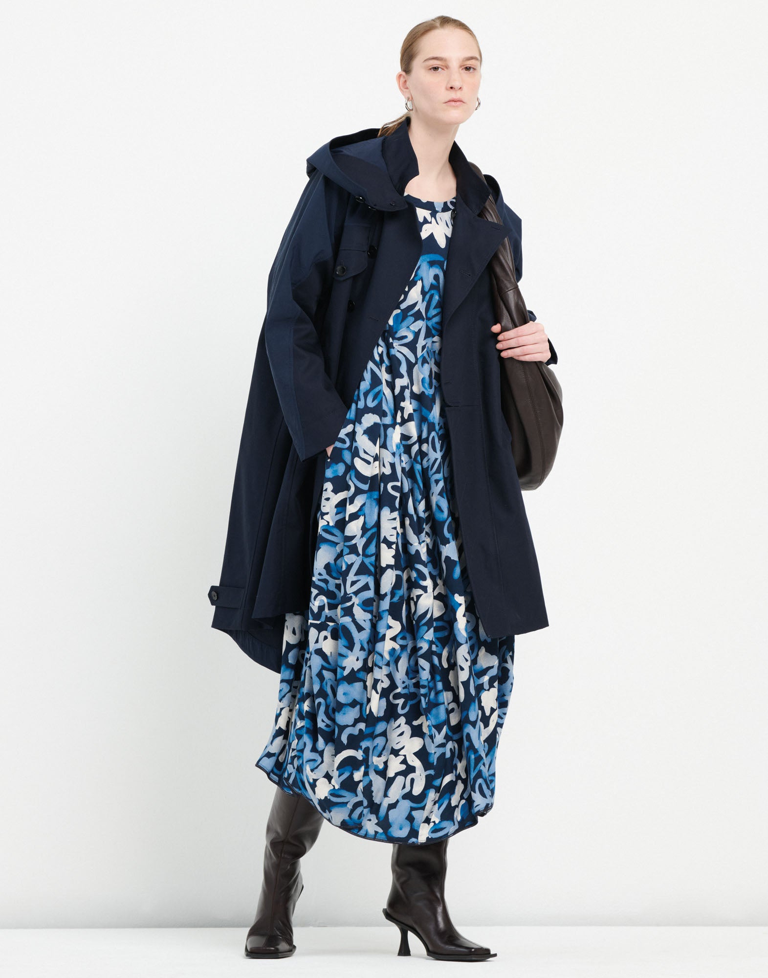 Front view: woman wears UPGRADE parka with integrated hood in dark blue, paired with a patterned blue dress and tall dark boots.
