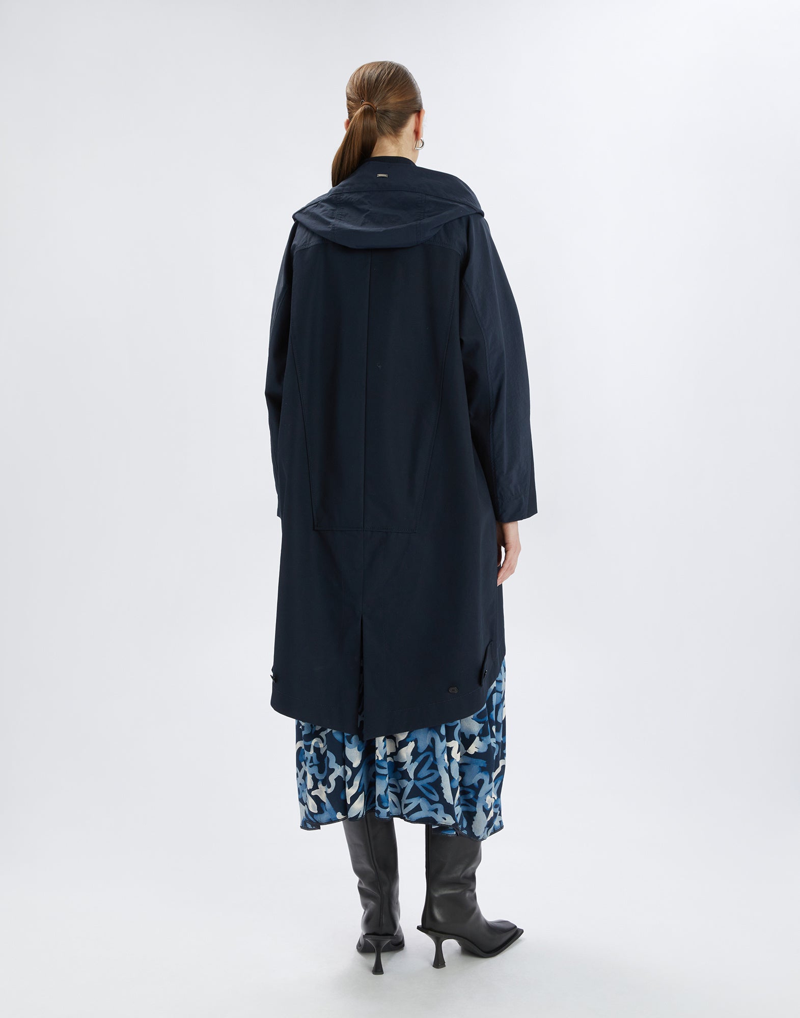 Rückansicht einer Frau, die den UPGRADE Parka trägt. Der oversized, dunkelblaue Parka mit Kapuze bedeckt ein gemustertes Kleid und wird mit Stiefeln kombiniert.