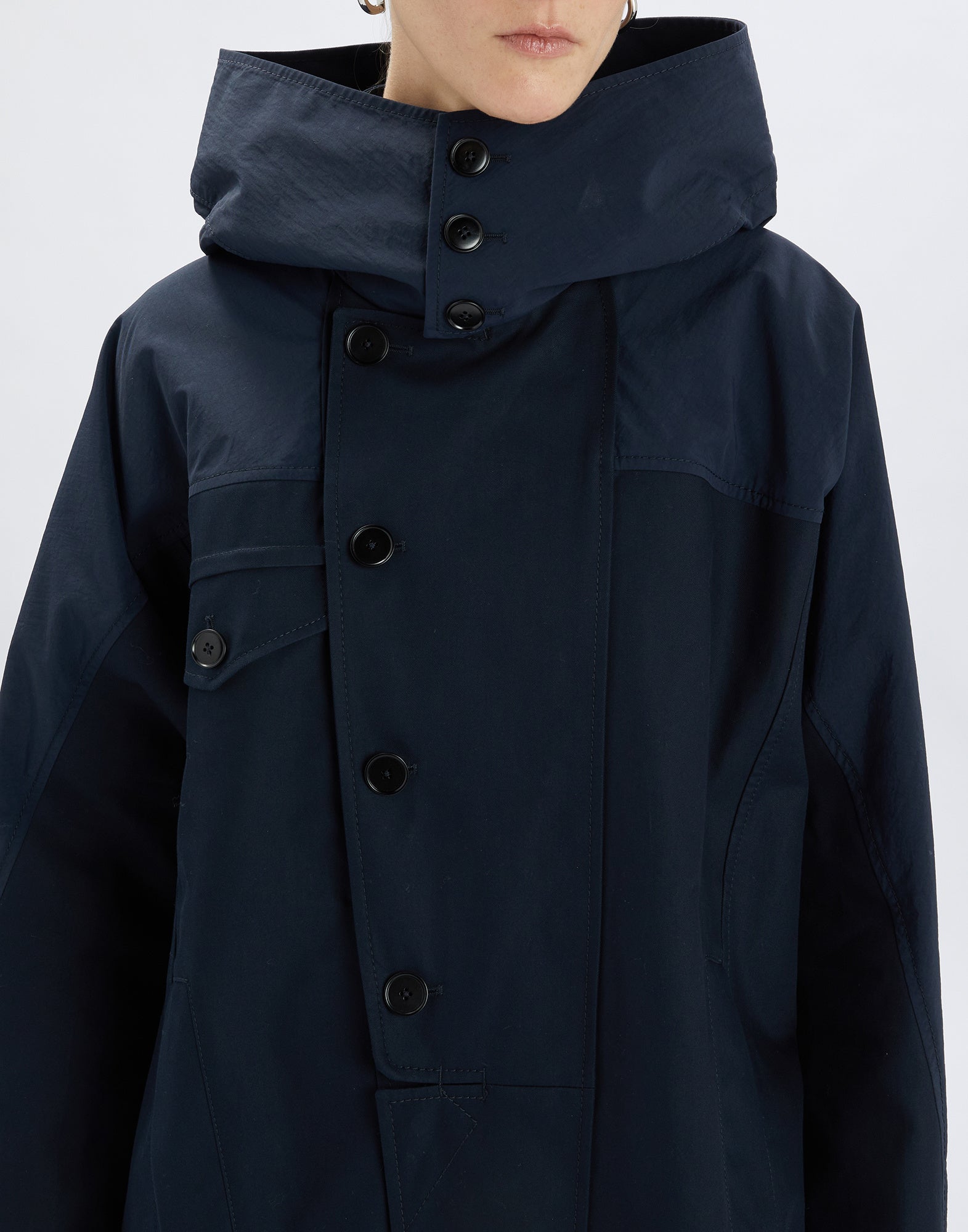 Frontalansicht des Parka UPGRADE, gezeigt von einer Person, mit hohem, geknöpftem Kragen und Kapuze, mattes, dunkelblaues Gewebe mit Taschen und Knopfleiste.