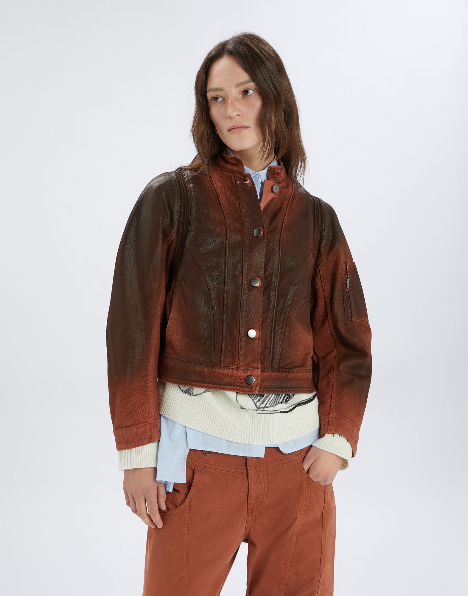 Front view of a woman wearing the TENDRIL jacket: boxy cut, brown-red leather-like look, stand collar, metal buttons, visible seams and zip pocket on the left sleeve.
