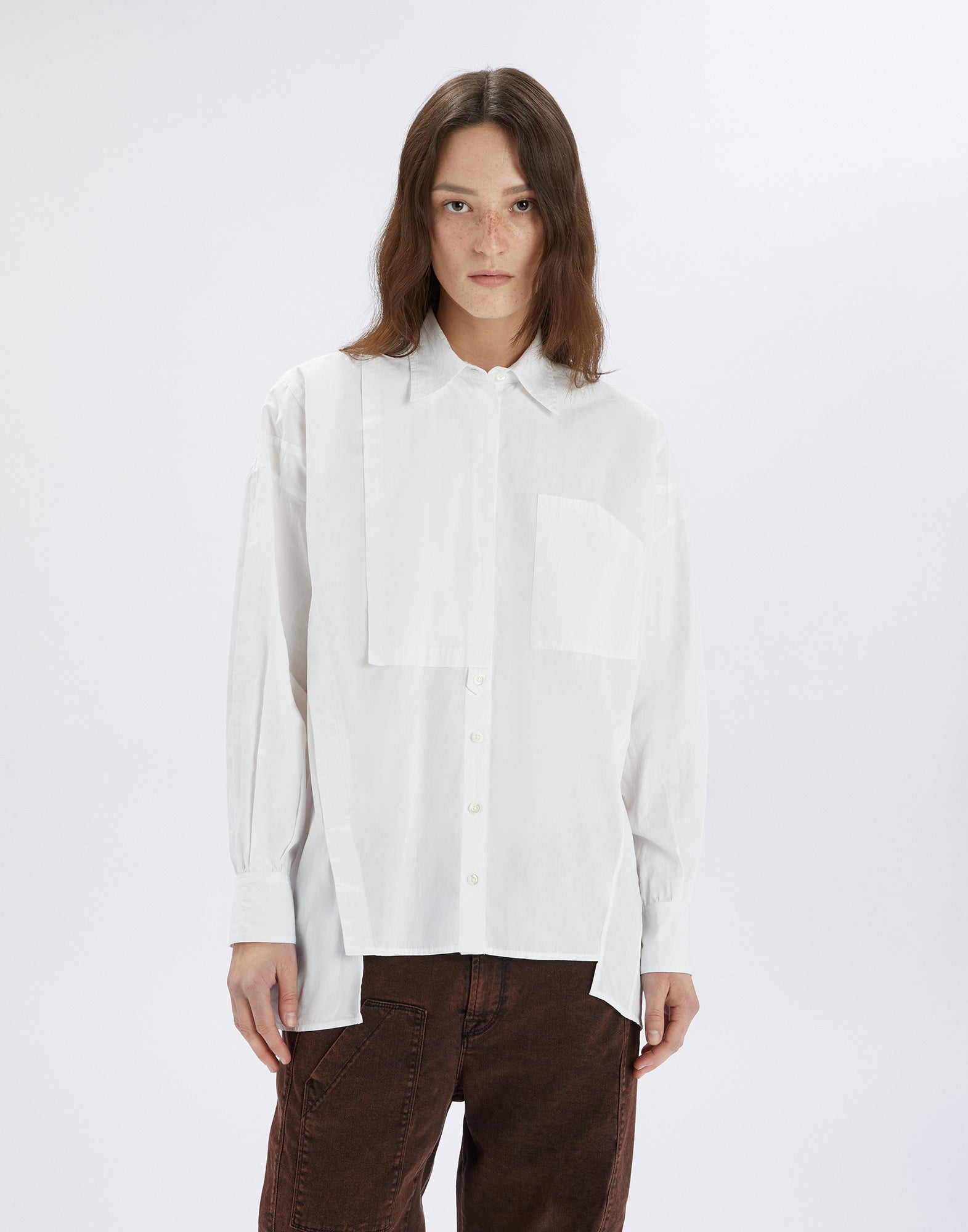 Front view: person wears the white DEADLINE shirt with loose fit, asymmetric front, chest pocket, and long sleeves, paired with brown trousers.