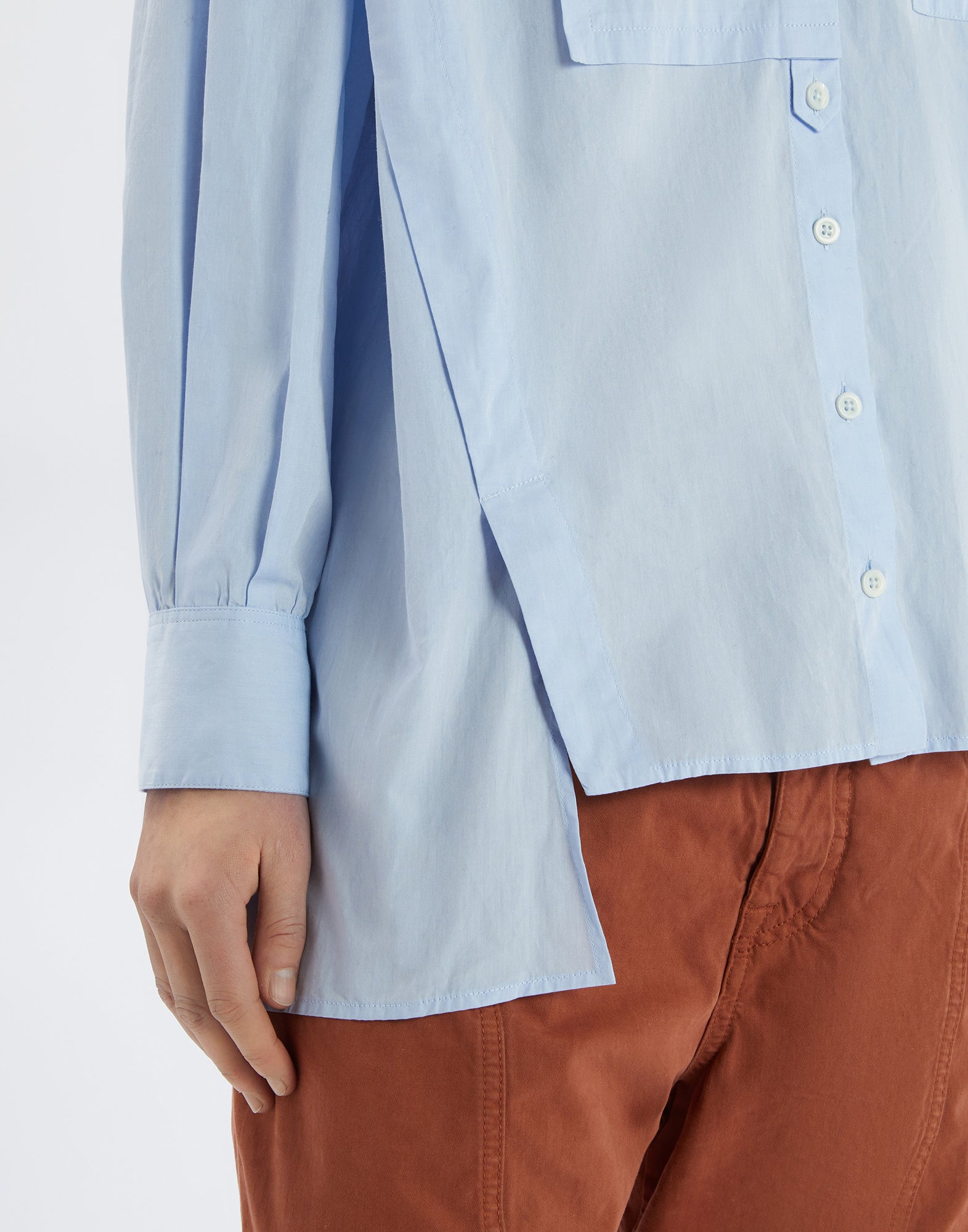 Detailansicht aus seitlicher Perspektive des DEADLINE Hemds in Hellblau, zeigt die lange Manschette, weiße Knöpfe und die asymmetrisch geschnittene Front über einer terrakottafarbenen Hose.
