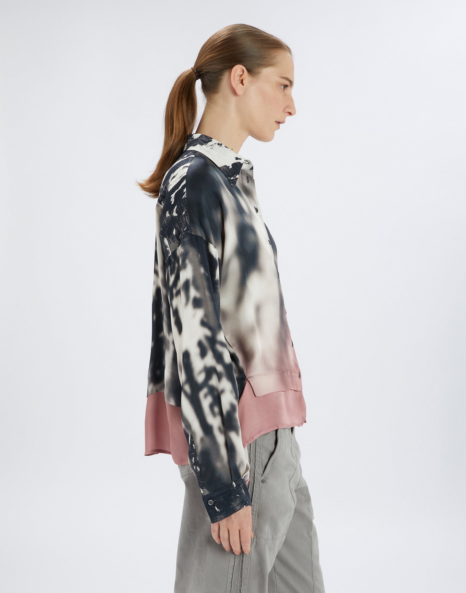 Side view of a person wearing the O-ZONE shirt. The shirt features a creative white-black contrast, softly transitioning into powder pink. Relaxed fit, turn-down collar, buttoned cuffs, and rounded hem are visible.