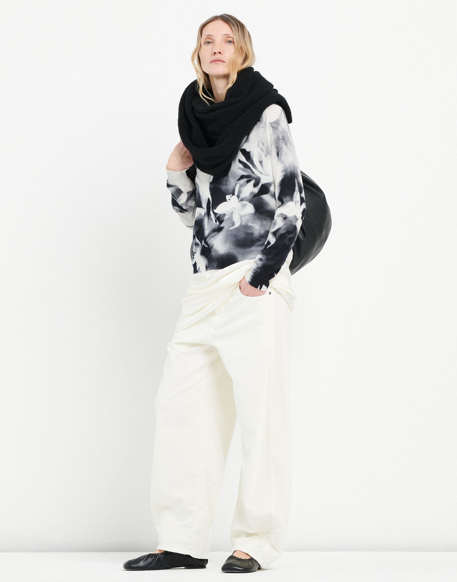 Frontalansicht einer Person, die den Pullover CECILIA mit floralem Schwarz-Weiß-Druck, weitem V-Ausschnitt und lockerer Passform trägt. Kombiniert mit weißer Hose und schwarzem Schal.
