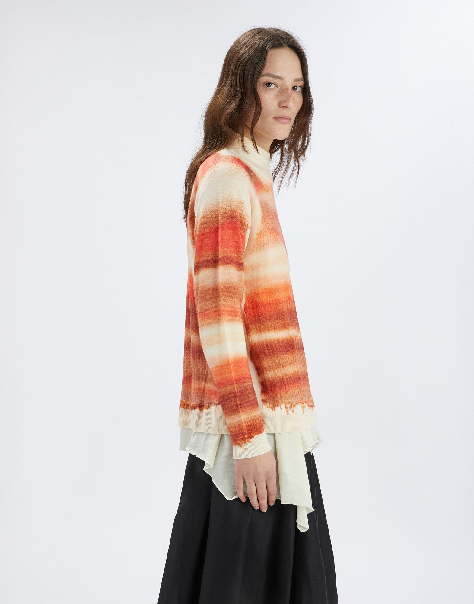 Seitenansicht einer Frau, die den Merinowollpullover RAMSEY trägt. Der Pullover zeigt einen Streifenprint in Orangetönen auf cremefarbenem Grund und hat Rippenbündchen.