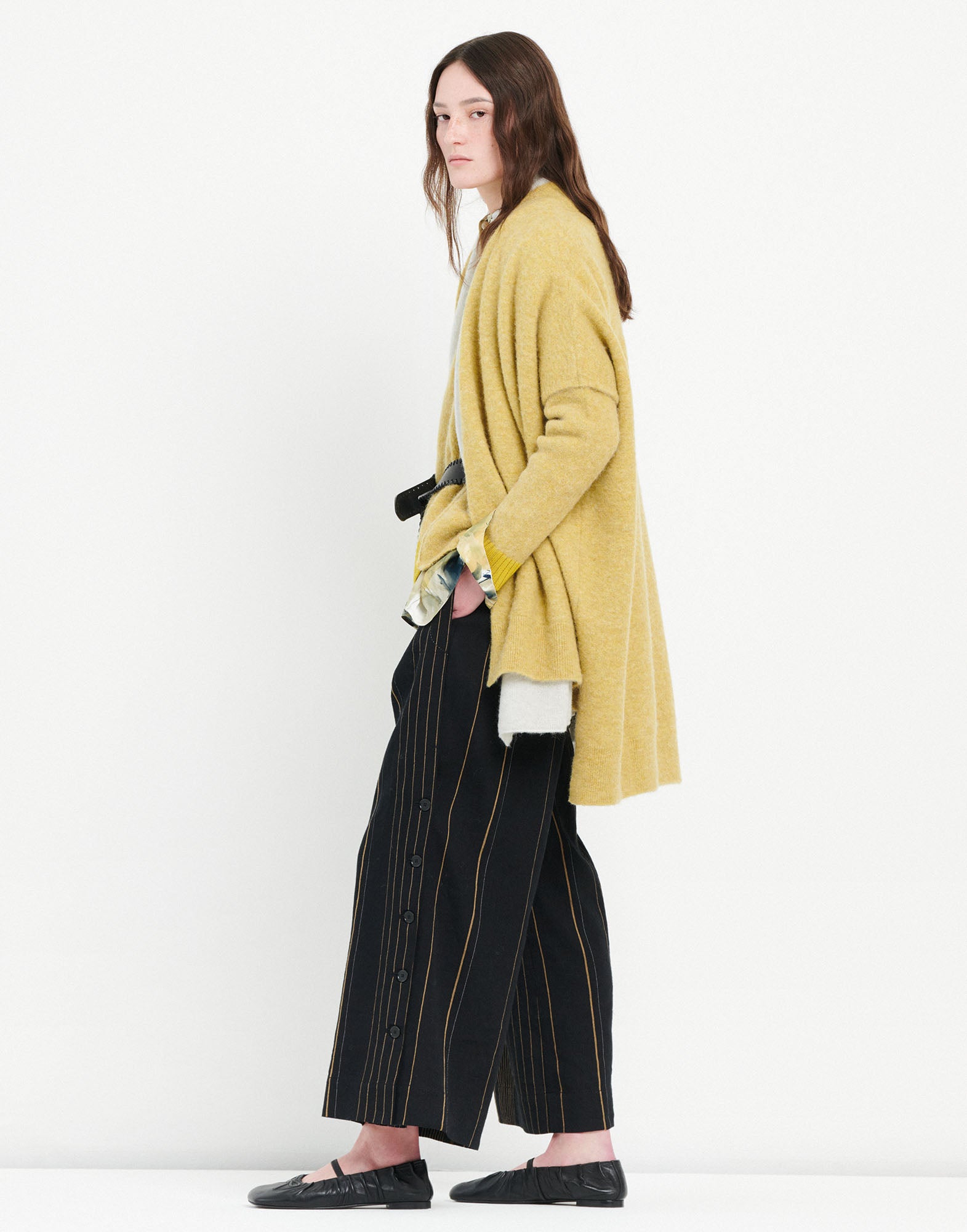 Seitenansicht des Umhangs PRUDENCE, aus flauschig wirkender, warmer Wolle und Alpaka, locker über einer Bluse und Hose getragen, offene Front, kurze Ärmel, gelbliche Farbe.