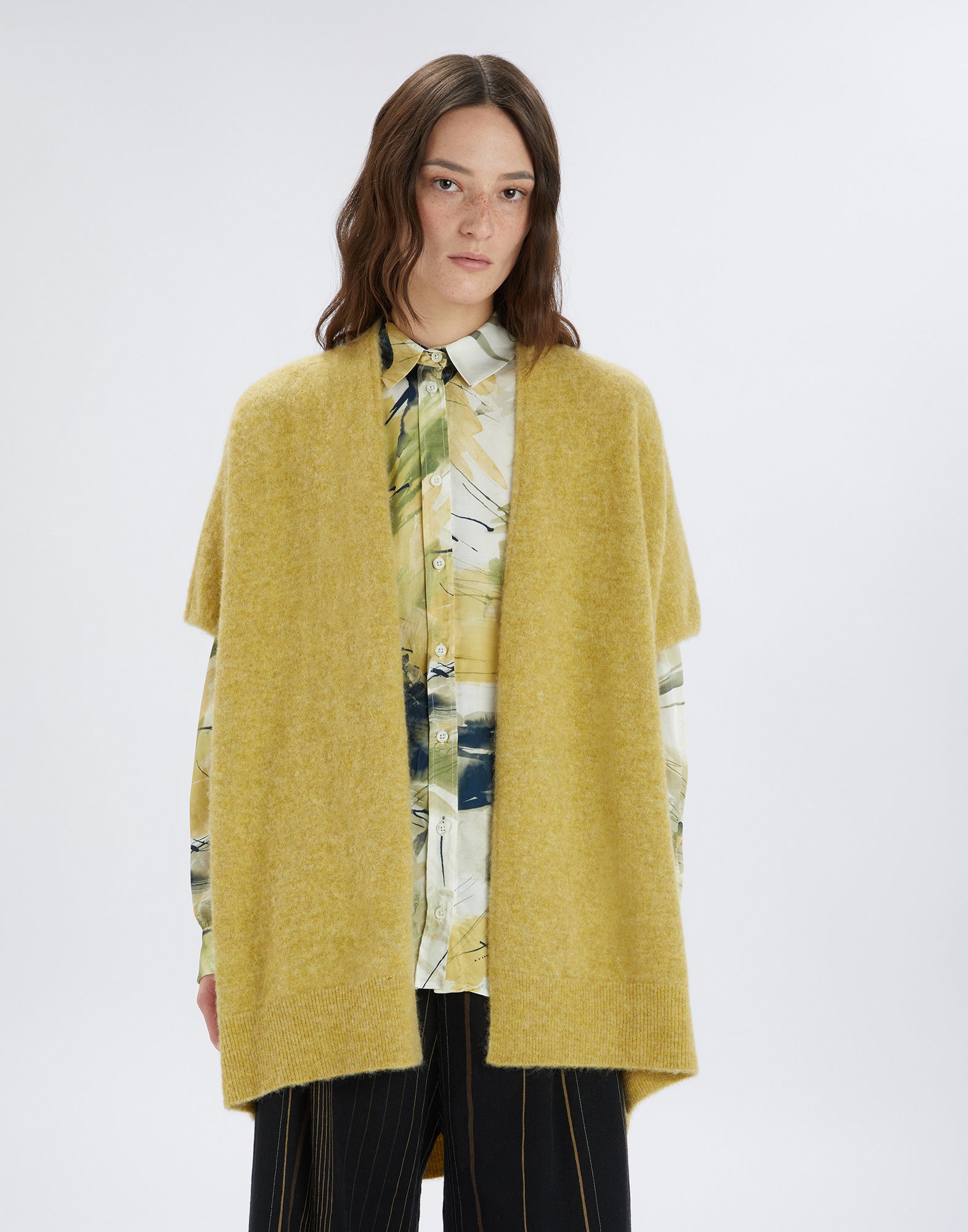 Front view: A woman wears the PRUDENCE cape made of yellow melange wool and alpaca, open front, with short sleeves and side slits over shirt and pants.