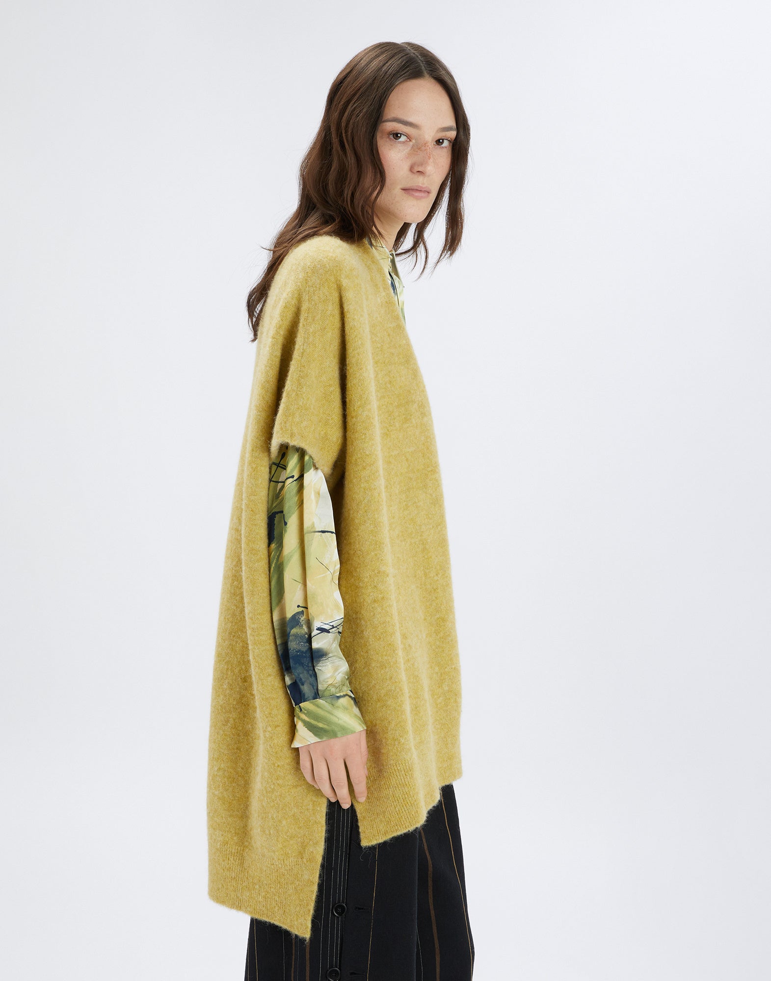 Side view: A woman wears the PRUDENCE cape in yellow melange wool and alpaca, draped loosely with short sleeves and side slits, over a patterned shirt.