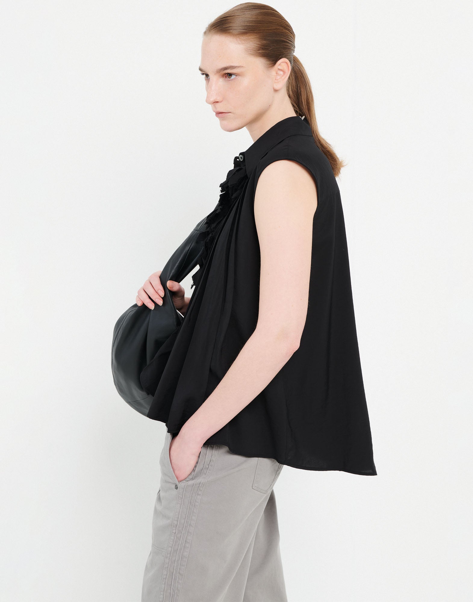 Seitenansicht eines Models, das die ärmellose Bluse DOODLE in Schwarz trägt; mit Reverskragen, unregelmäßigen Falten und abnehmbarer floraler Applikation am Kragen.