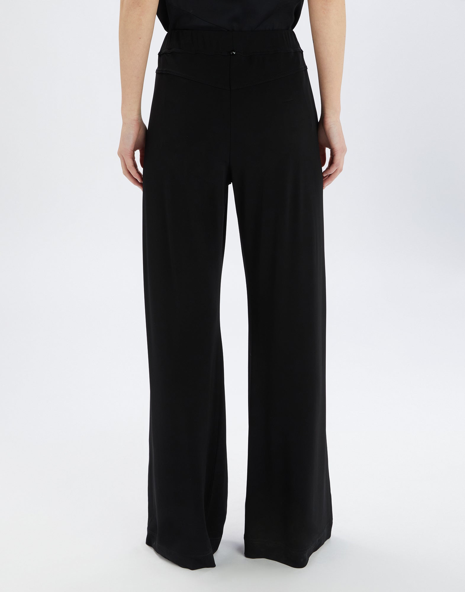 Rückansicht einer Person, die die SLIP-OVER Hose aus schwarzem Viskosejersey trägt. Weit geschnittenes Bein, elastischer Bund, leicht glänzender Stoff.