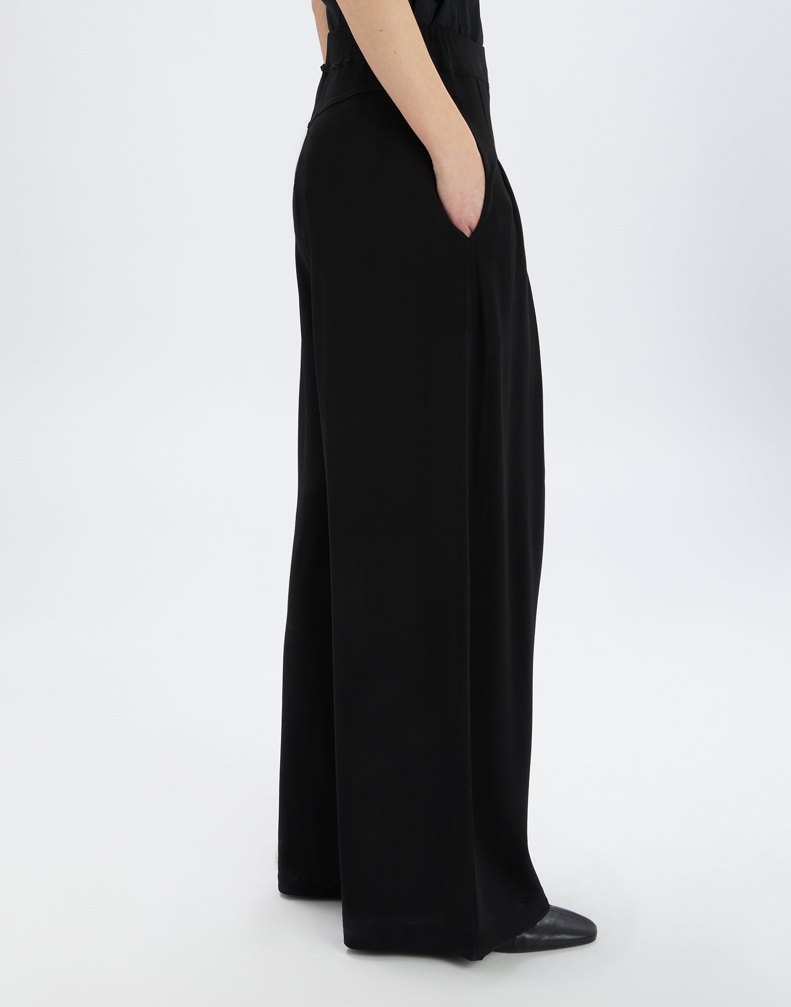 Seitenansicht der SLIP-OVER Hose, getragen von einer Person mit einer Hand in der seitlichen Eingrifftasche. Der Schnitt ist weit und aus schwarzem, fließendem Viskosejersey gefertigt.