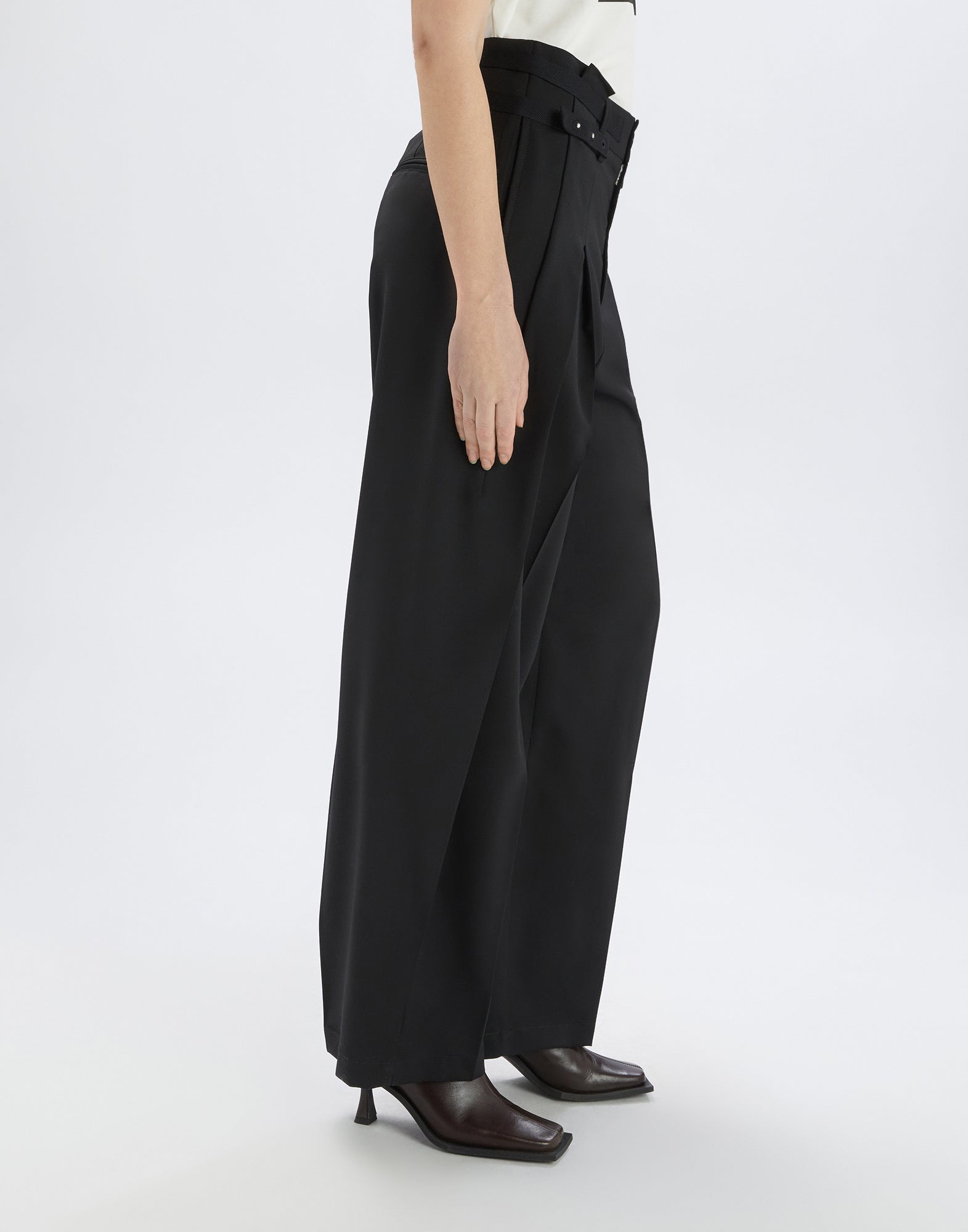 Side view of NEVERLAND pants in black extra-fine wool, relaxed fit, double front pleats, modern waist belt with rivets, worn with heeled shoes.