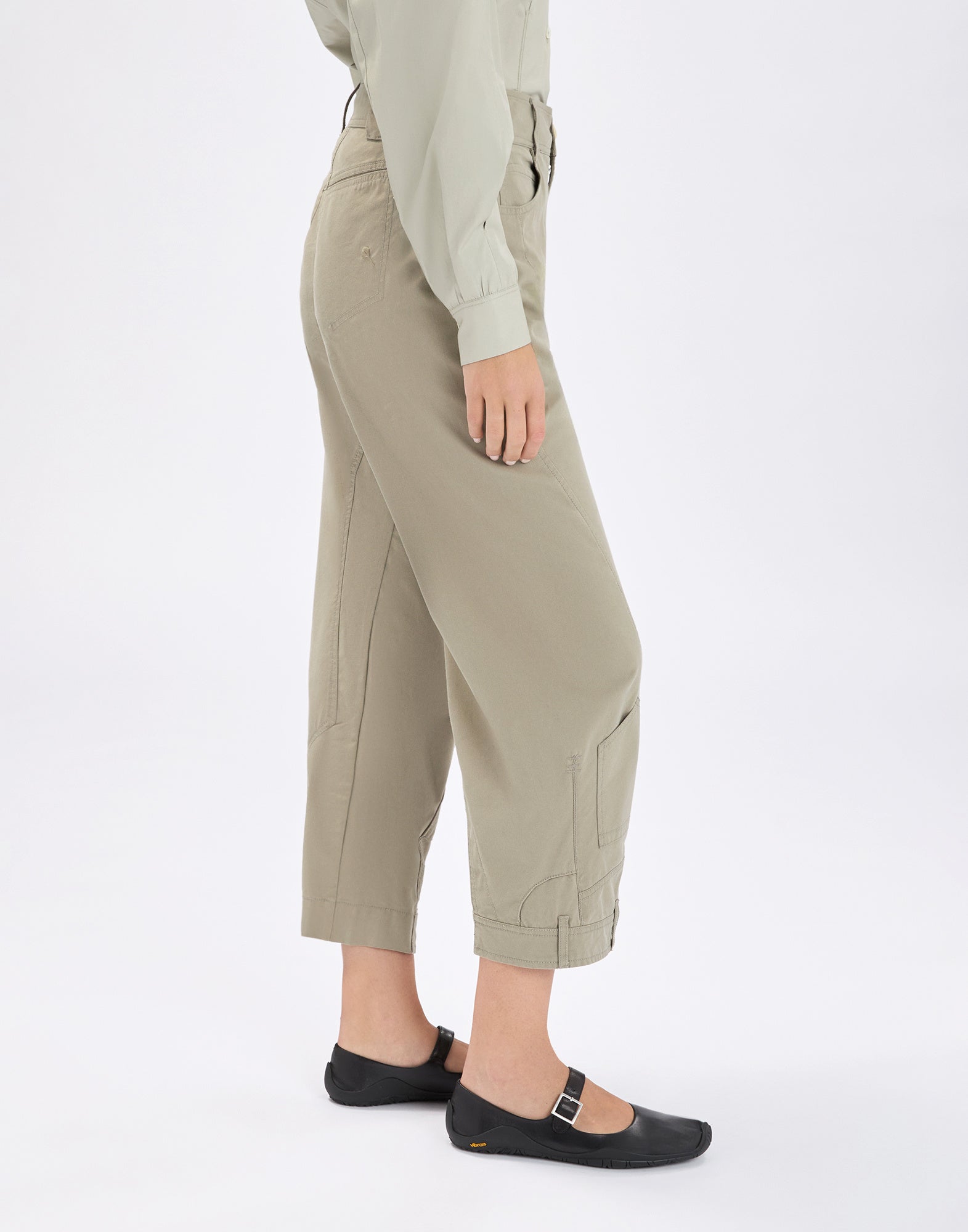 Side view: person wearing khaki LOLA pants with wide ankle-length leg, visible seams, patch pockets and belt-loop-style tabs at the hem, paired with a blouse and black flats.