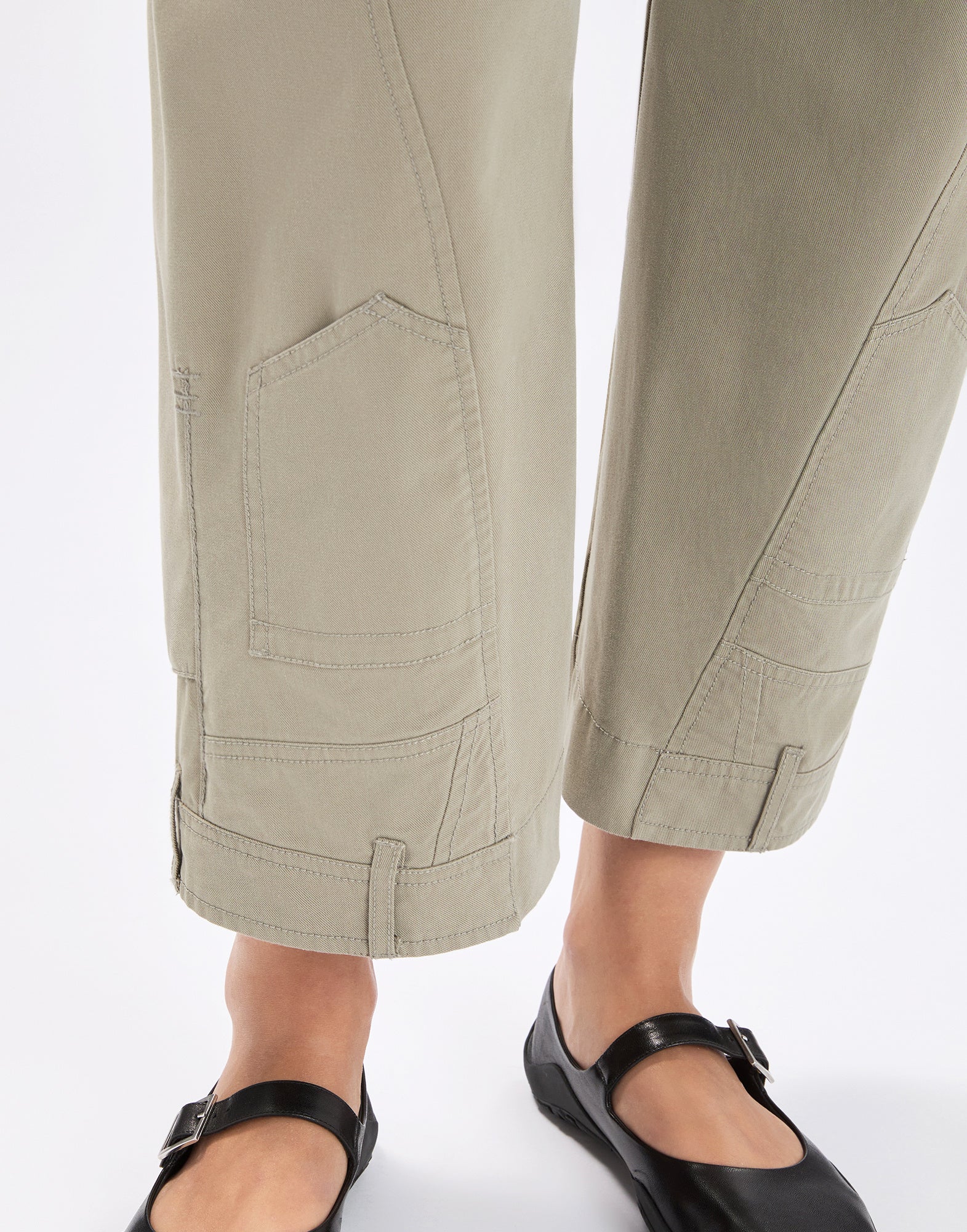 Detail view: lower leg section of the LOLA pants from the front, beige wide legs with upside-down patch pockets, visible seams and belt-loop-style tabs at the hem, paired with black Mary Jane shoes.