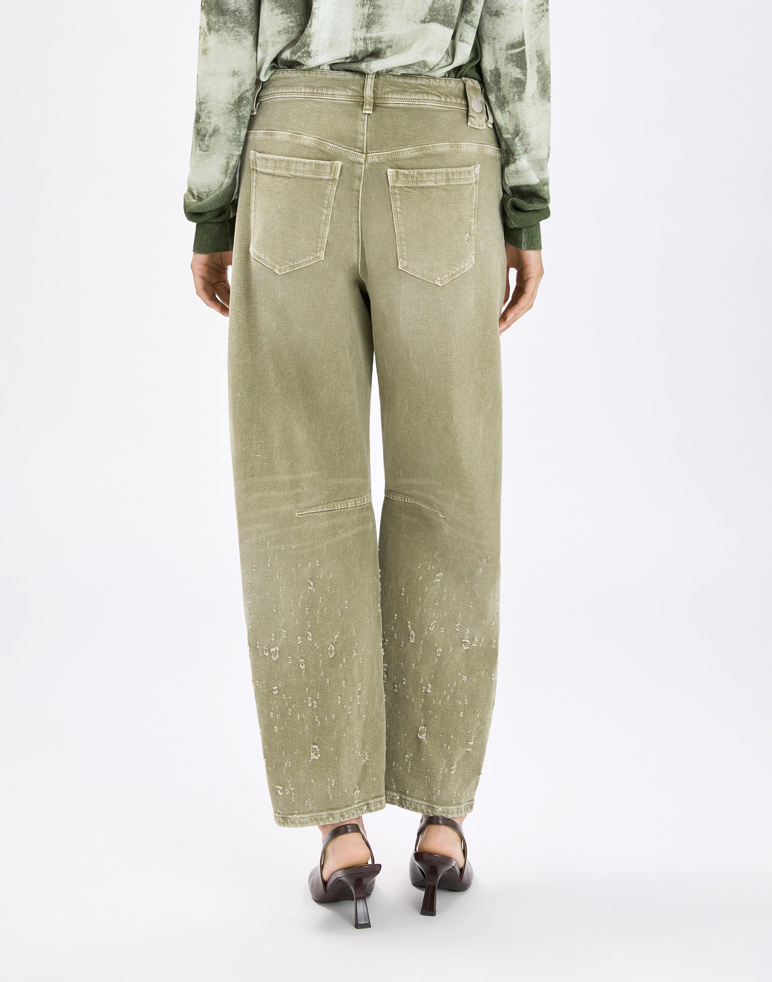 Back view: person wearing HAPPILY pants in olive green vintage look, with exposed seams, two patch back pockets, wide leg and subtle distressing effects, paired with heeled sandals.