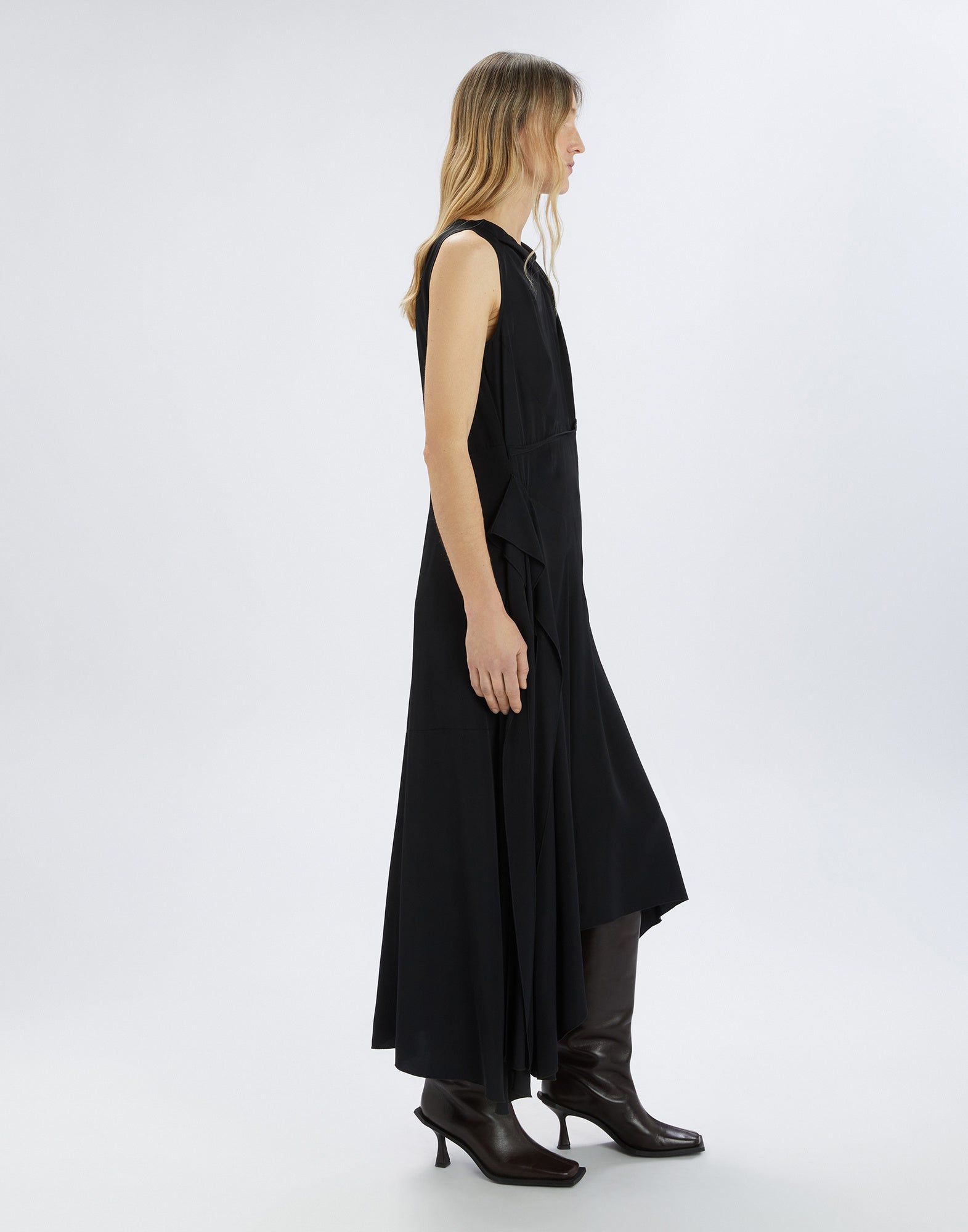 Seitenansicht eines ärmellosen, schwarzen SOUND OFF Kleids mit asymmetrischem Saum, fließendem Stoff und seitlicher Drapierung, getragen von einer Frau mit Stiefeln.