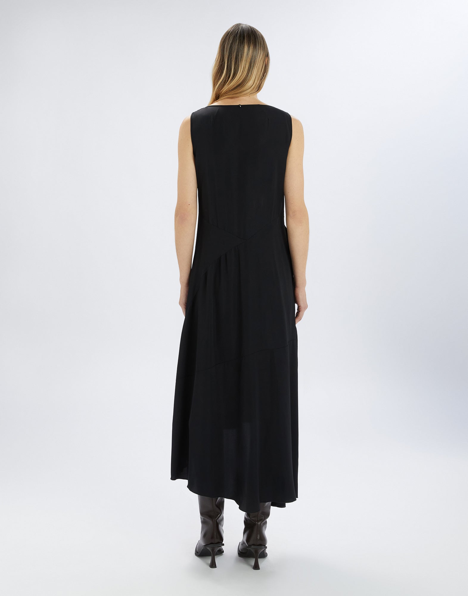 Rückansicht des ärmellosen Kleides SOUND OFF in Schwarz, das locker über die Hüfte fällt und eine asymmetrische Saumlinie aufweist. Model trägt dunkle Stiefel.