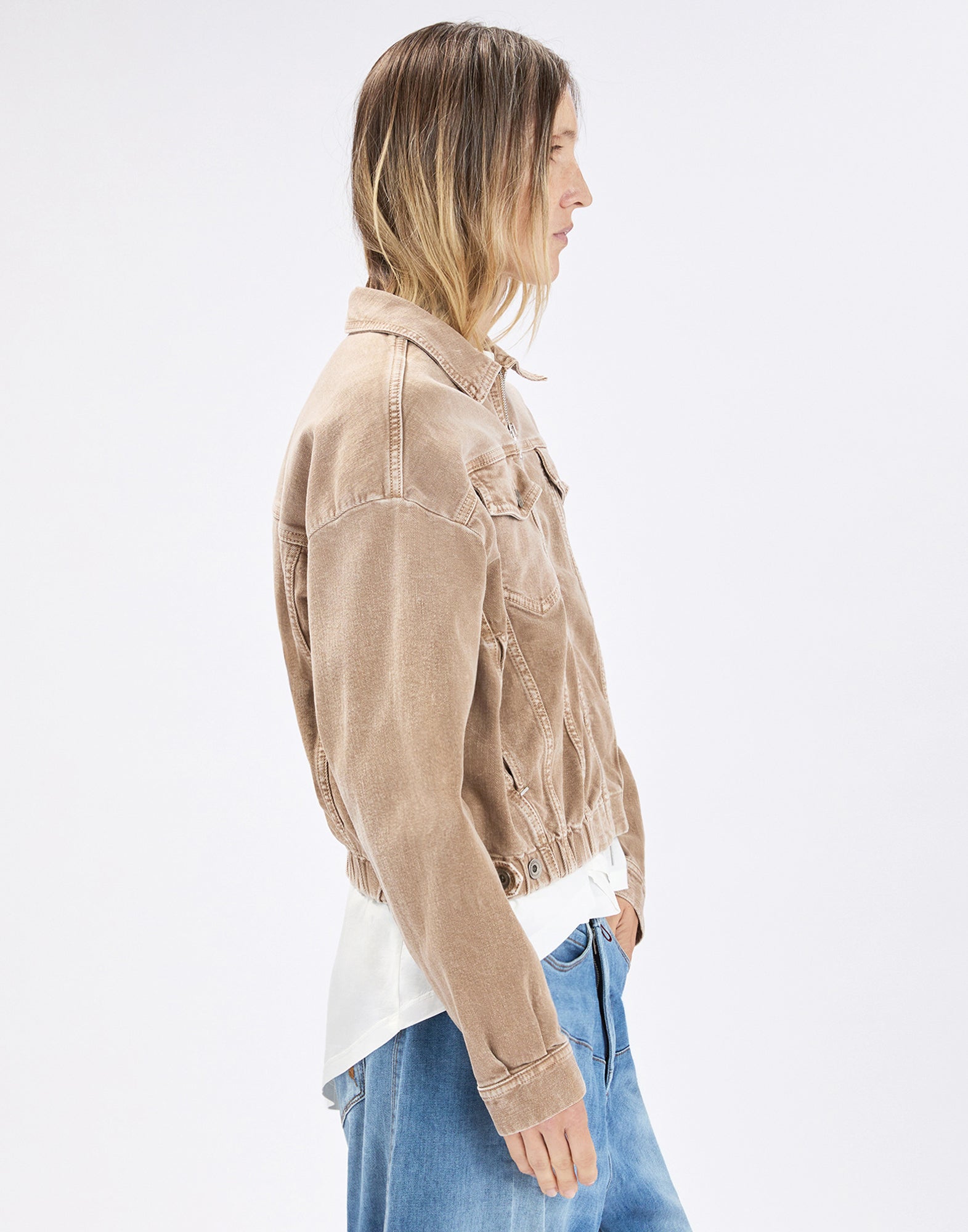 Seitenansicht: Person trägt die beige Denim-Bomberjacke COMPACT mit umgeschlagenem Kragen, Brusttaschen mit Patte, elastischem Bund und Knopf-Manschetten über weißem Shirt und blauer Jeans.