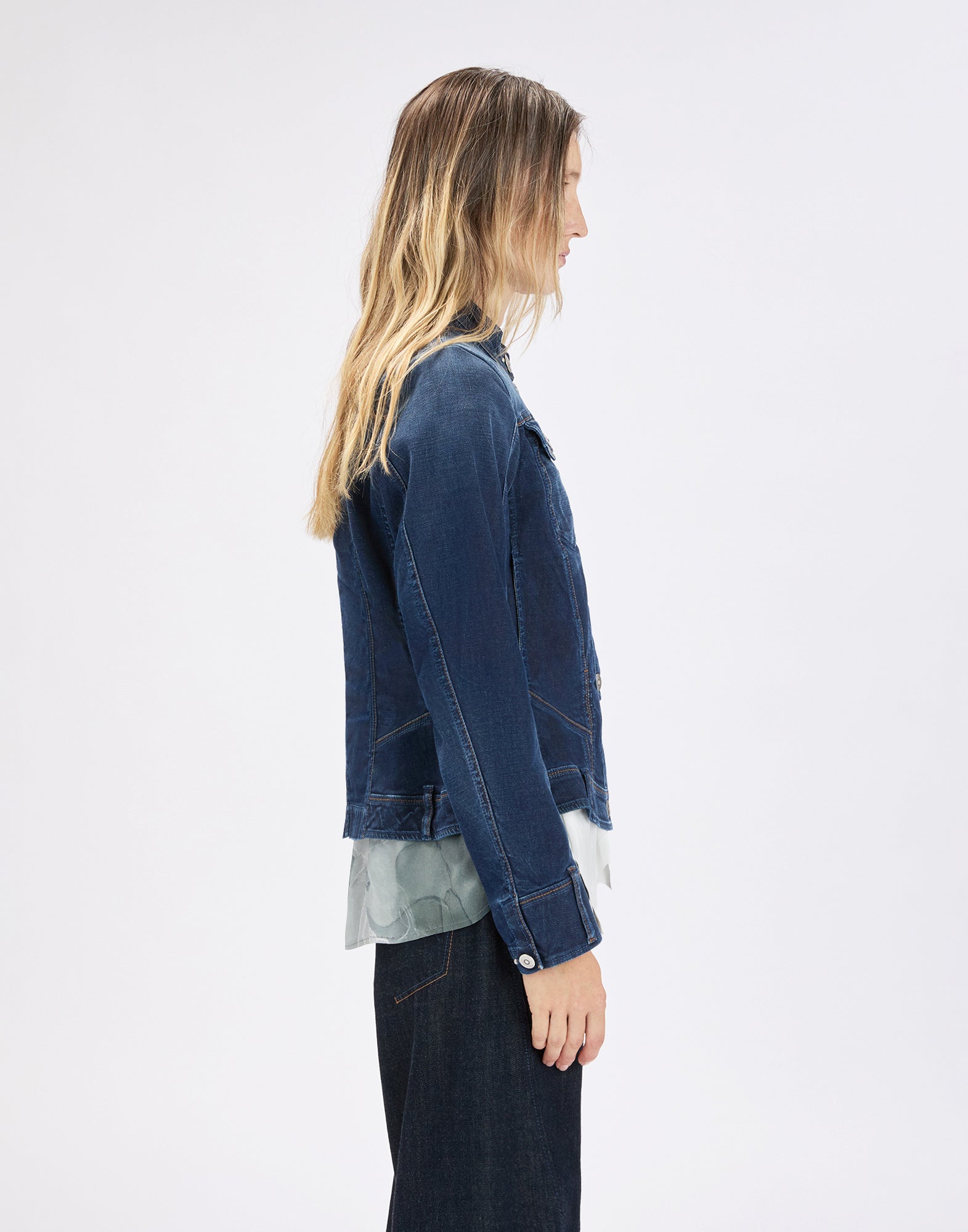 Side view: person wearing the dark blue AHOY denim jacket in a slim fit with visible stitching, chest pockets and buttoned cuffs, over a light top and dark trousers.