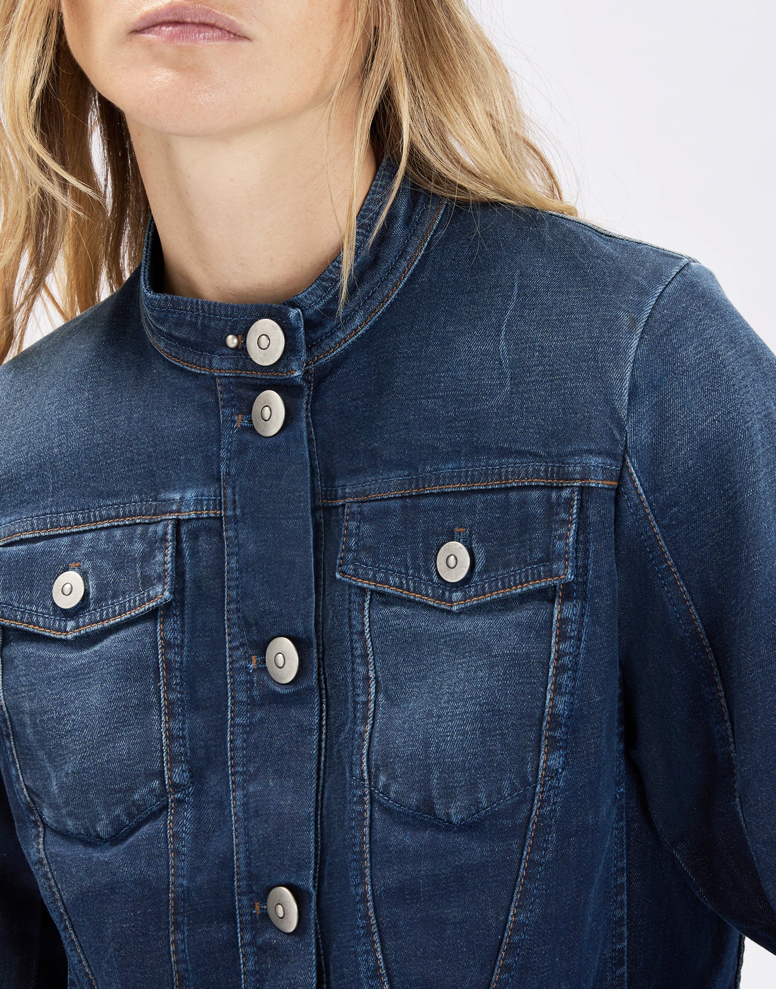 Detail view, front view: model wearing the dark blue, fitted AHOY denim jacket with high stand-up collar, silver metal buttons, flap chest pockets and pronounced corset-style seams.