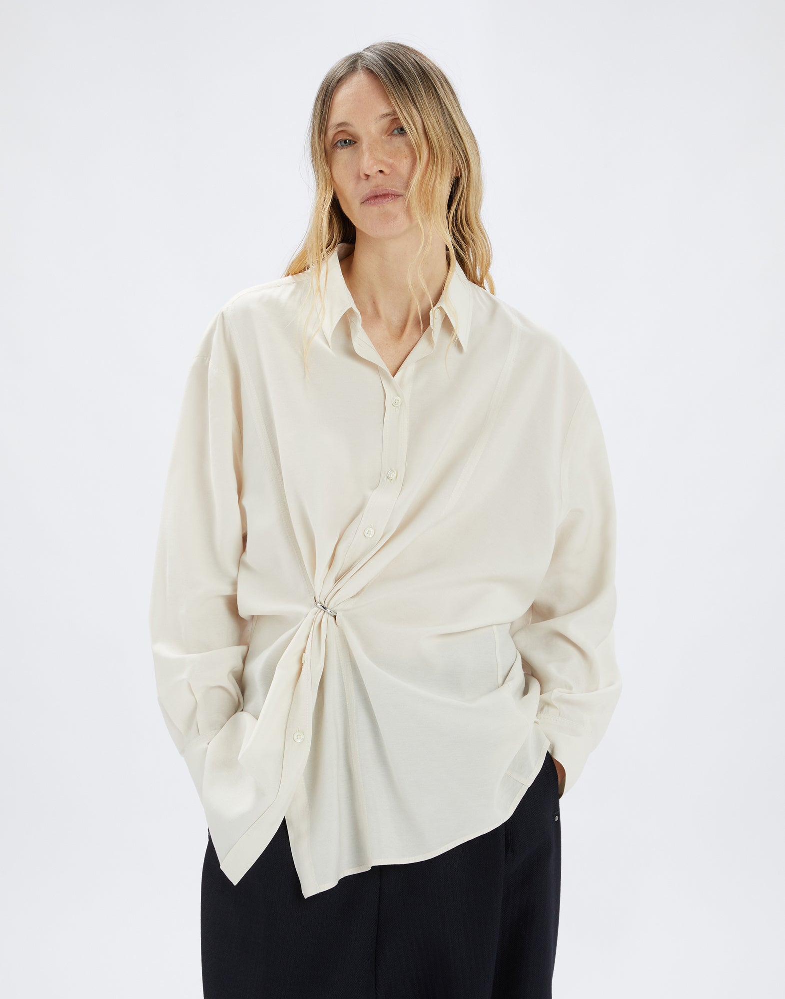Front view: woman wears SKYLARK blouse in light color with loose fit, classic collar, central button placket, and prominent ring detail creating draping.