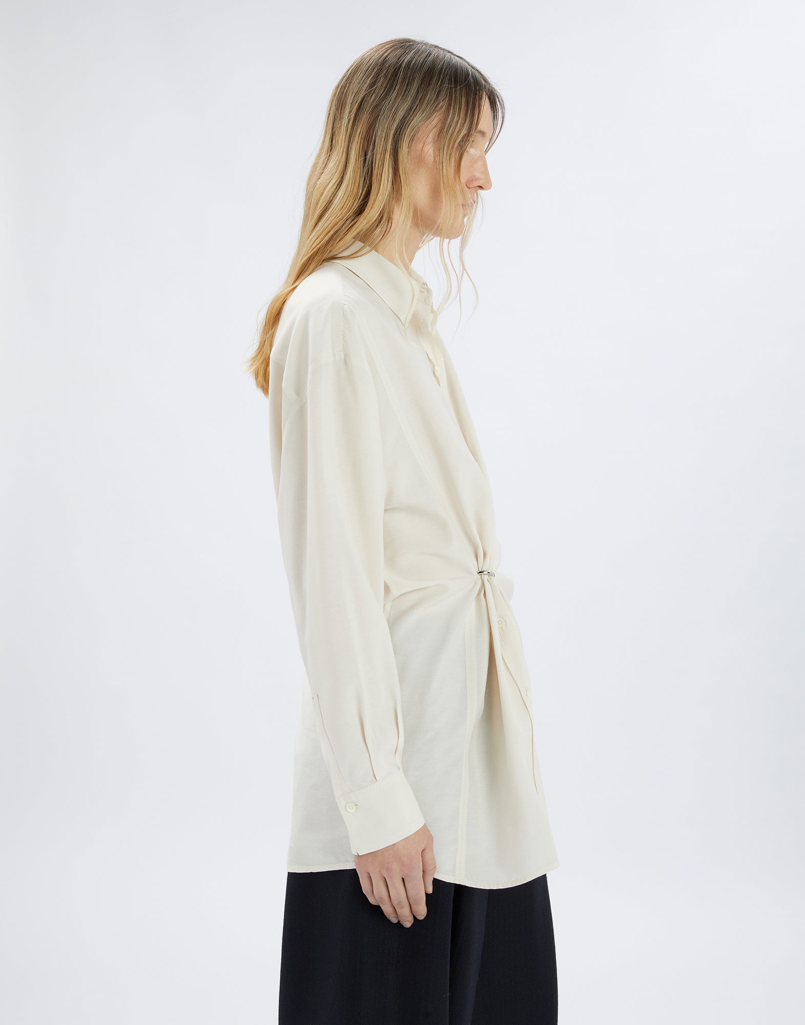 Side view of a person wearing the cream SKYLARK blouse; loose fit with turned-down collar, long button placket, and central D-ring for draping.