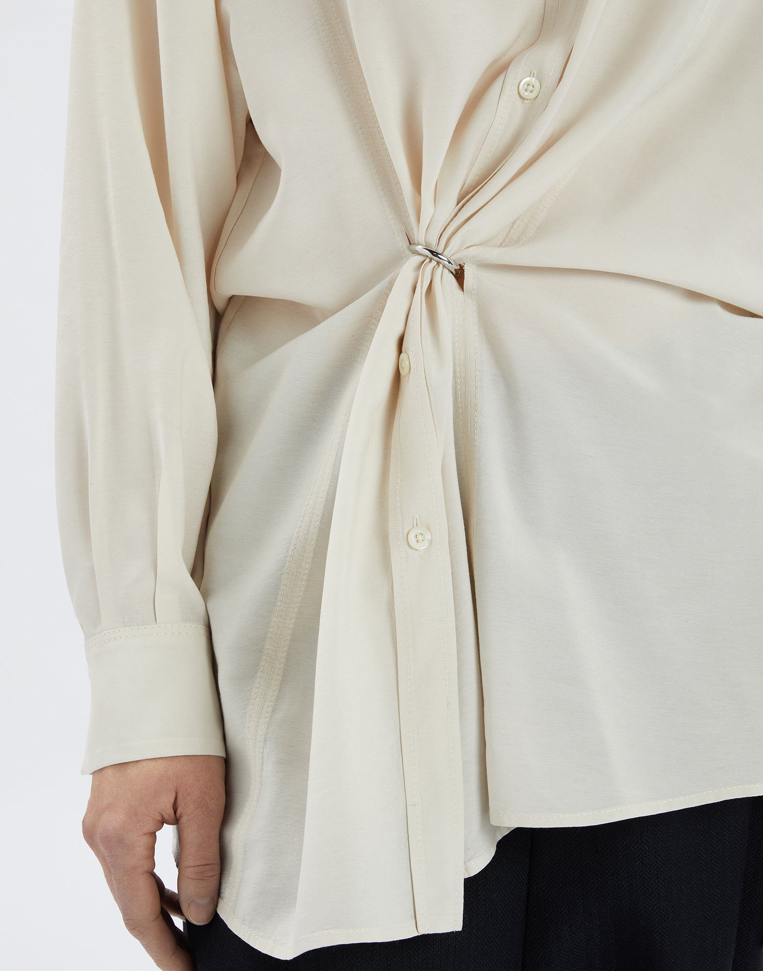 Close-up view of a person wearing the cream-colored SKYLARK blouse with central D-ring, elegant pleats, long sleeves, and visible buttons.