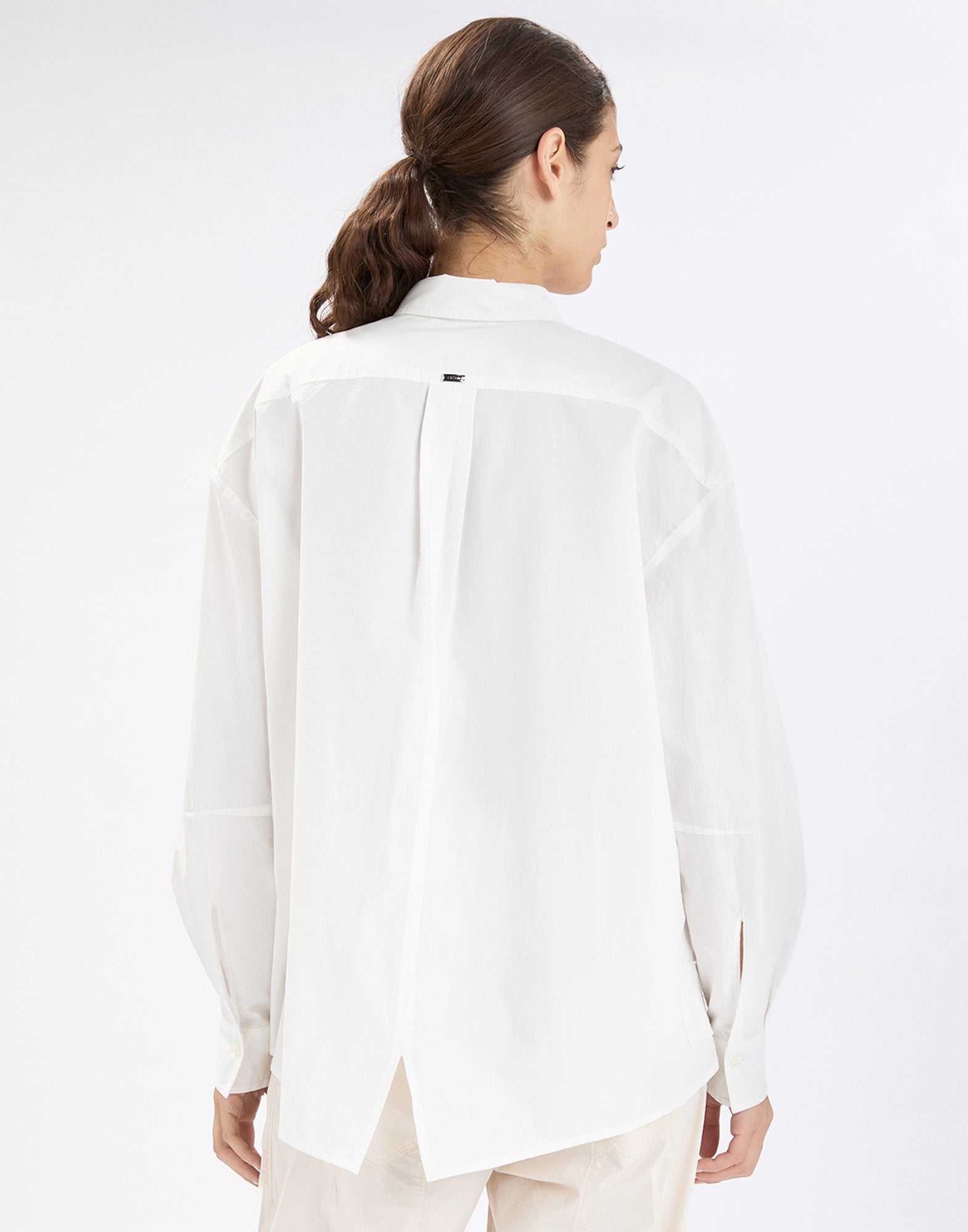 Rear view: Person wearing the white SAWYER shirt with a long, extended back, center box pleat, small back label, and slightly asymmetric, slit hem over light-colored pants.