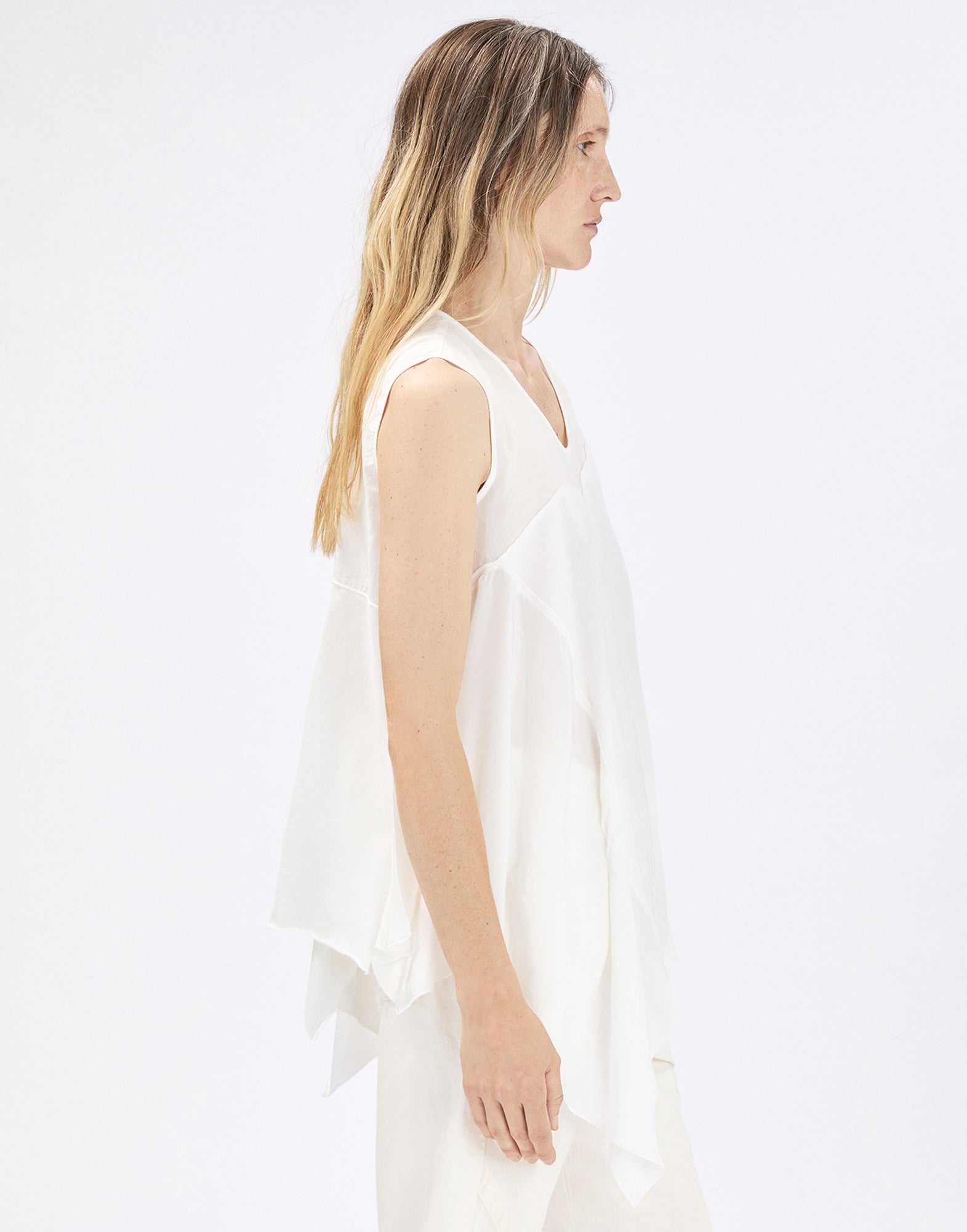 Side view: model wears sleeveless, loose-fitting white COMPASS top with V-neckline, asymmetric ruffle and irregular hem over light-colored trousers against a neutral background.