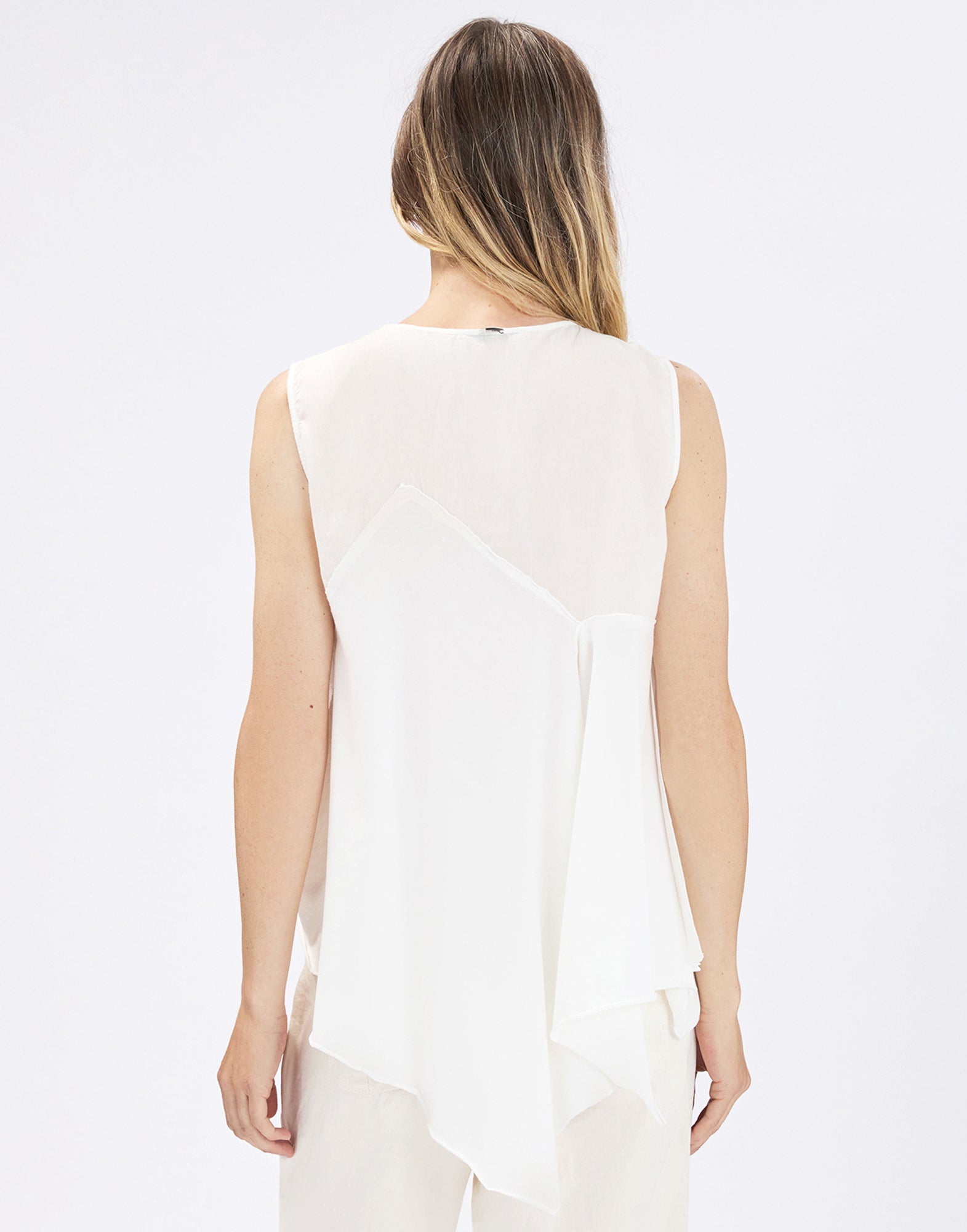 Rear view: Person wearing the sleeveless COMPASS top in cream with a softly draping asymmetric ruffle and irregular hem over light, wide-leg pants against a neutral background.