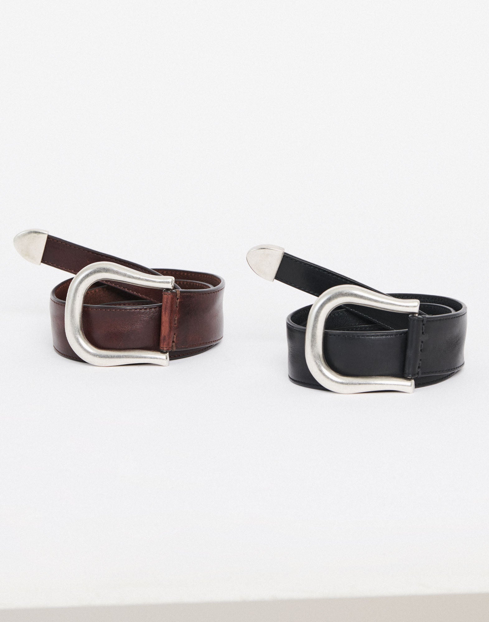 Front view of two SHORTY belts made of calf leather, one in dark brown and one in black, both featuring a large aged silver metal buckle.