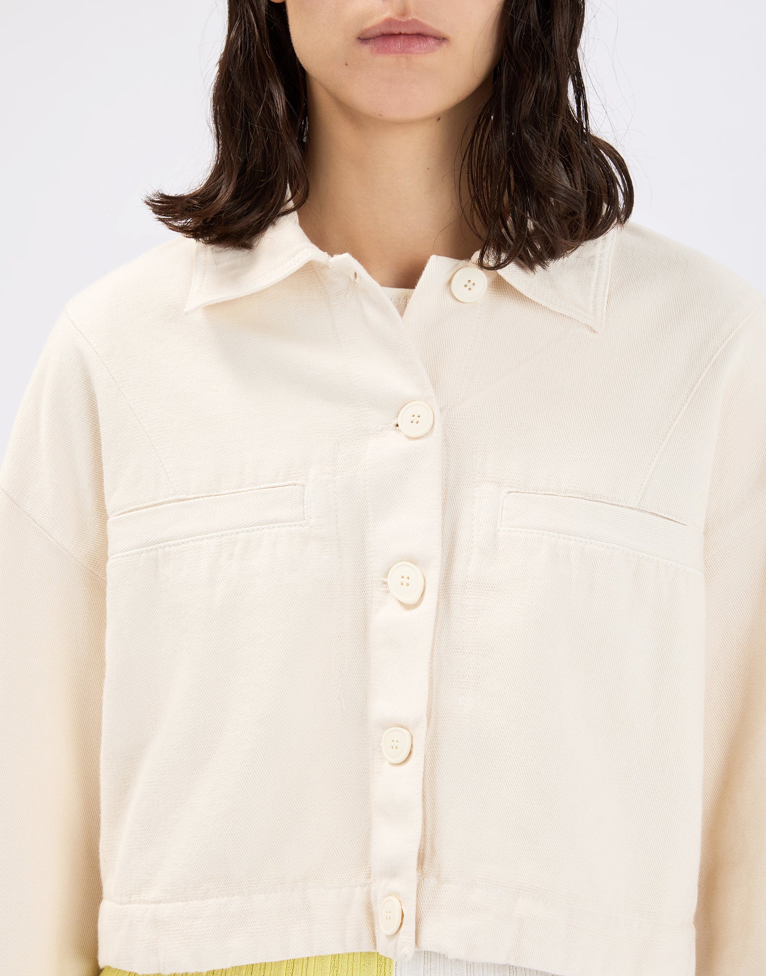 Detail view, front view: model wearing the cropped, cream-colored DEBUT denim jacket with shirt collar, full-length button placket, and two subtle chest pockets in a cotton-linen blend.
