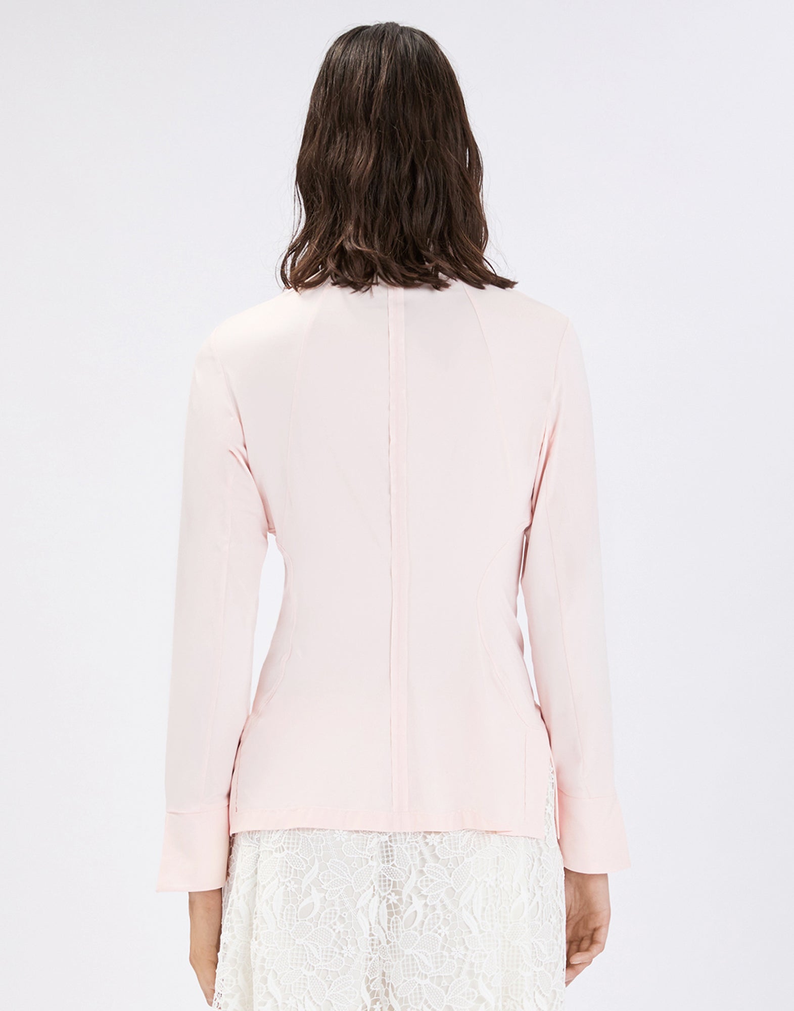Rear view: Person wears the form-fitting light pink INDEED shirt with long sleeves; visible panel seams accentuate the waist, paired with a white lace-trimmed skirt.