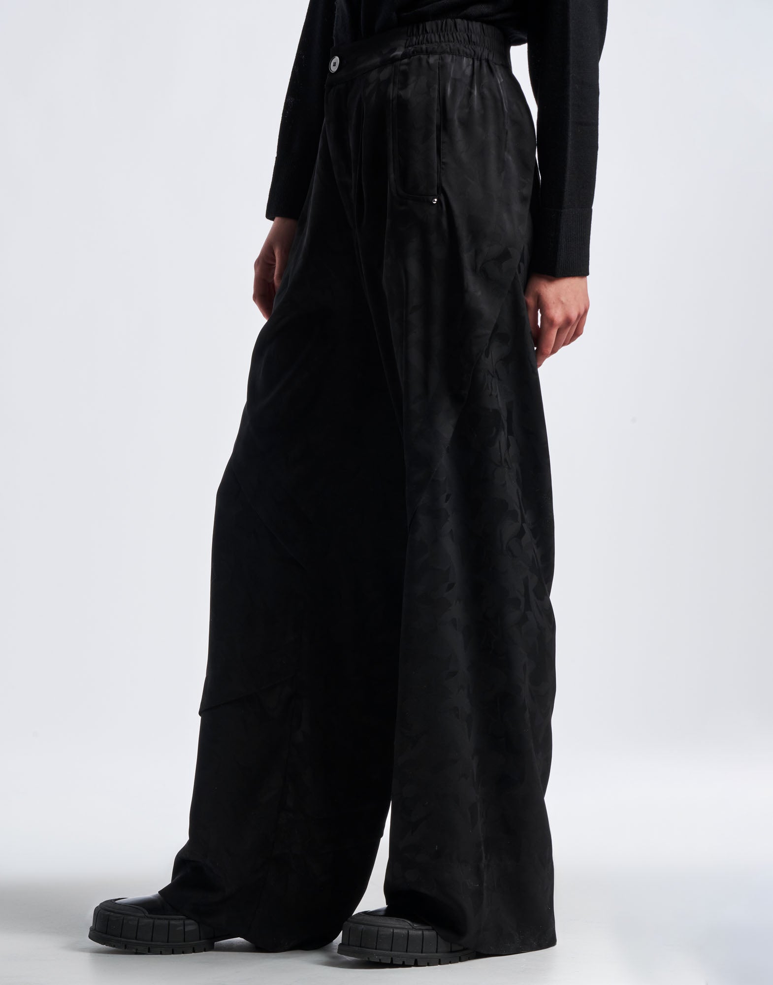 Side view of BORDERLINE pants in black technical satin with floral damask pattern, wide and loose-cut leg, and visible button at waistband.