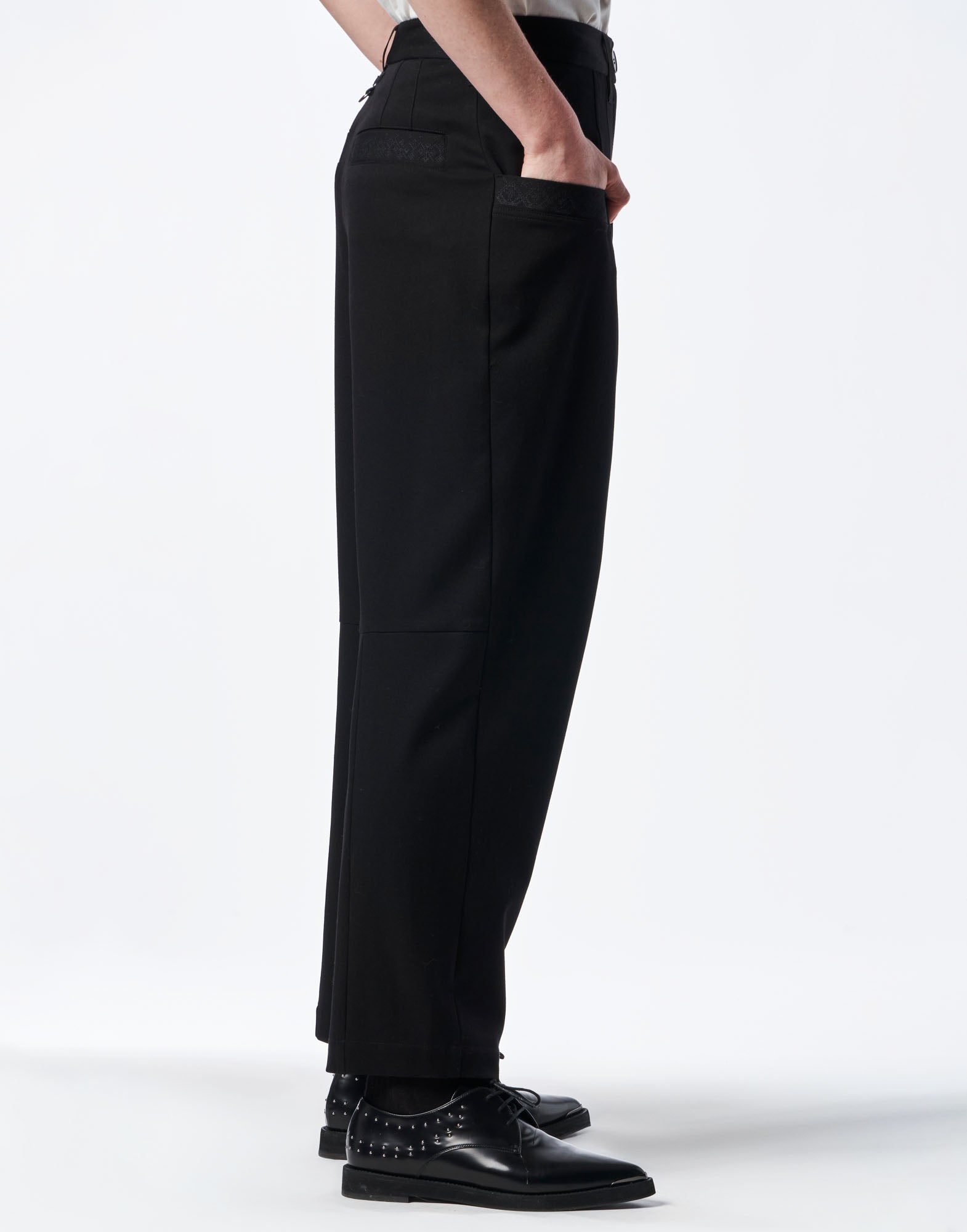 Side view of the black COMPLY pants worn, featuring visible belt loops, side welt pockets, and a tapered cut against a light background.